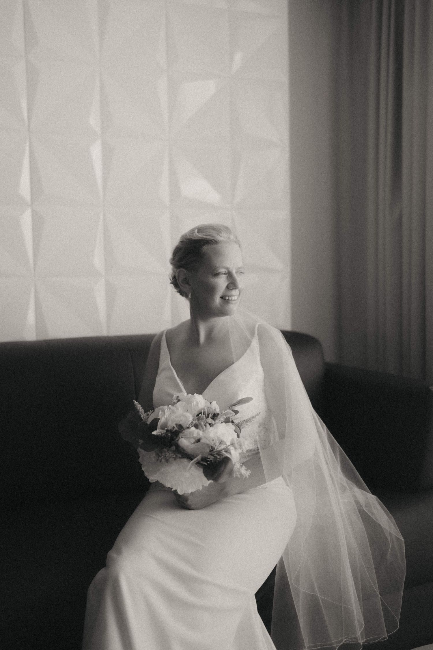 Bride in white dress and veil sitting by window in elegant black and white portrait series.