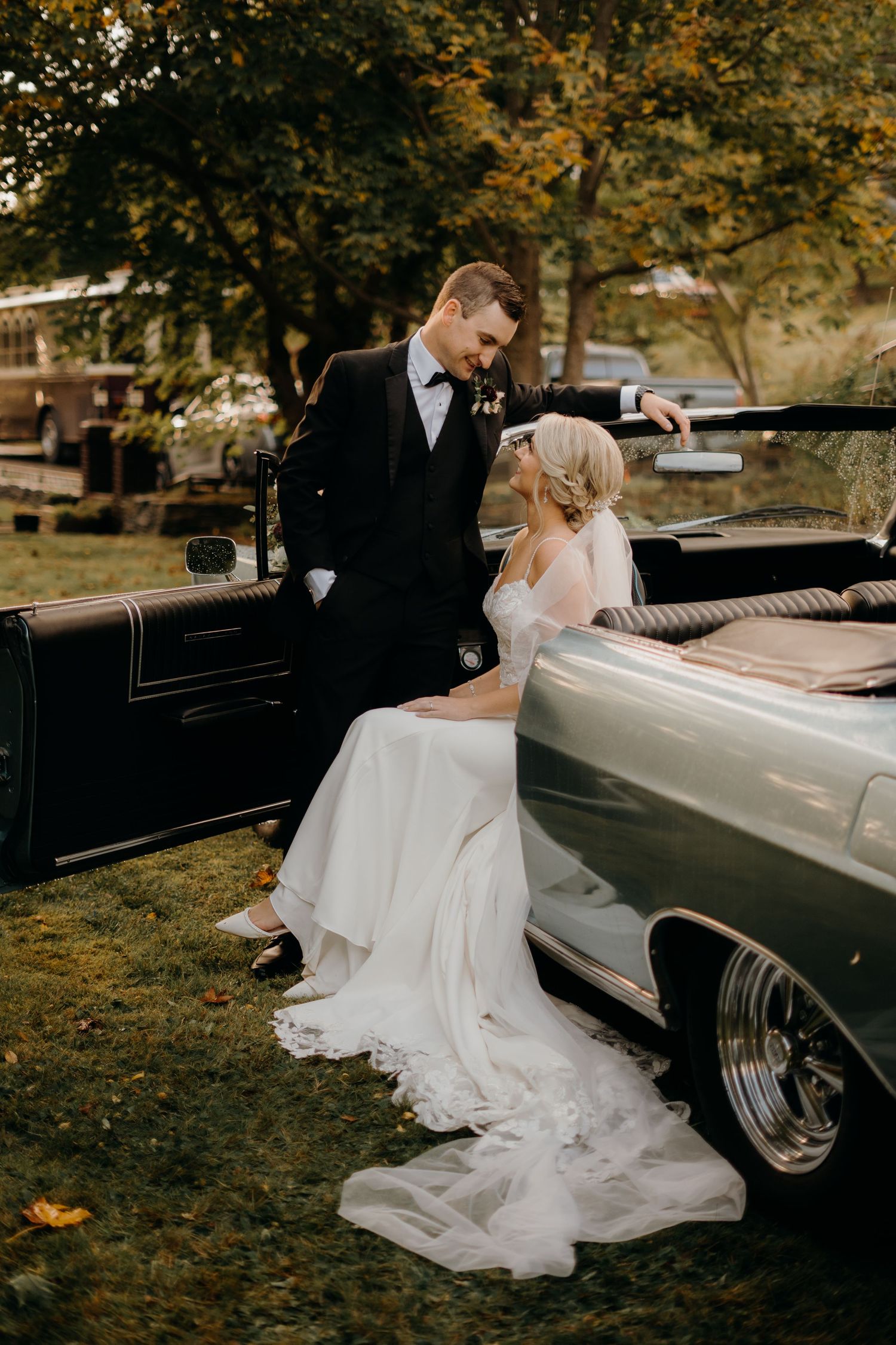 Elegant wedding couple poses by a classic vintage car surrounded by autumn leaves.
