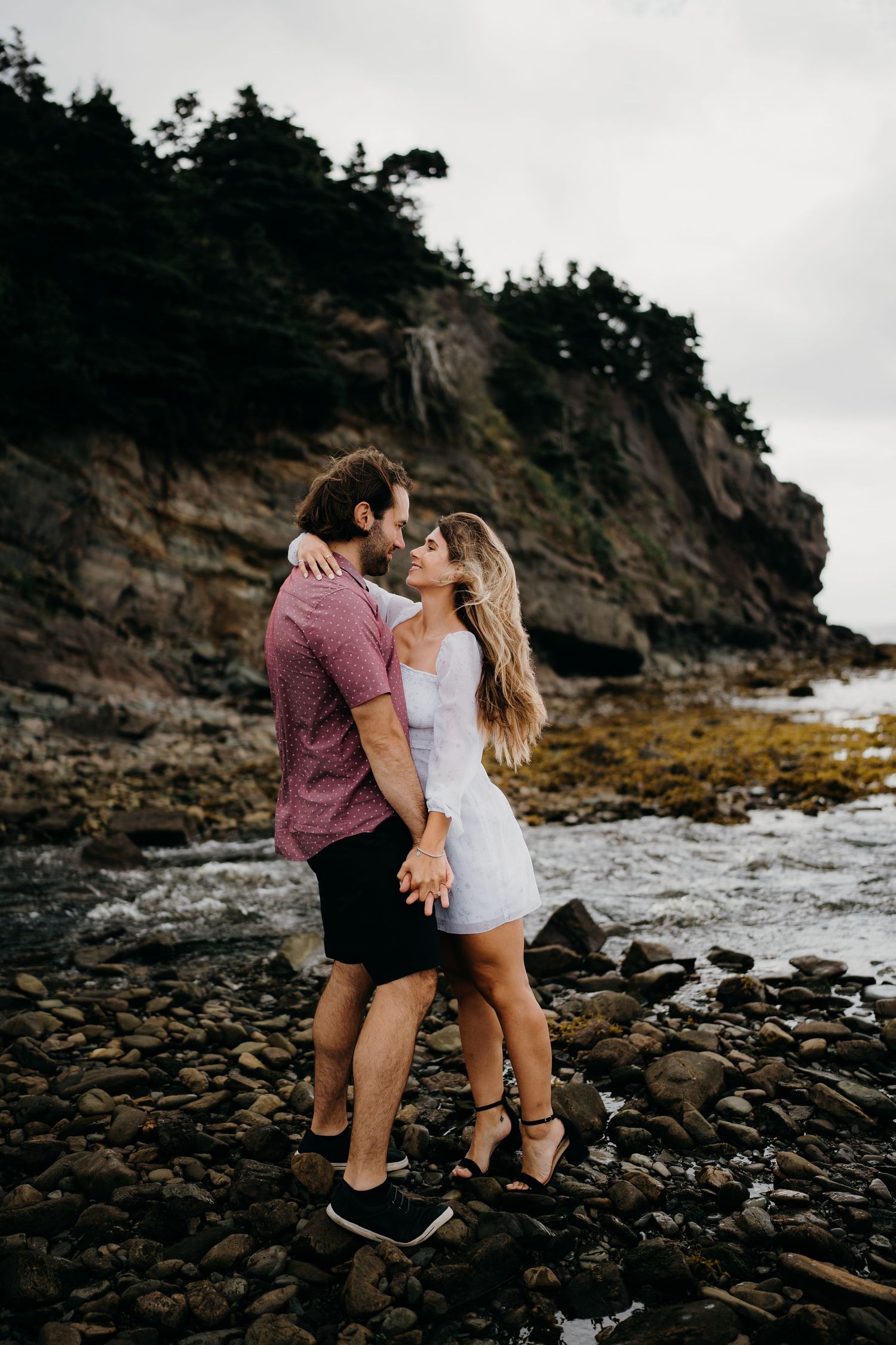 A romantic couple shares an intimate moment on a rocky beach coastline at sunset.