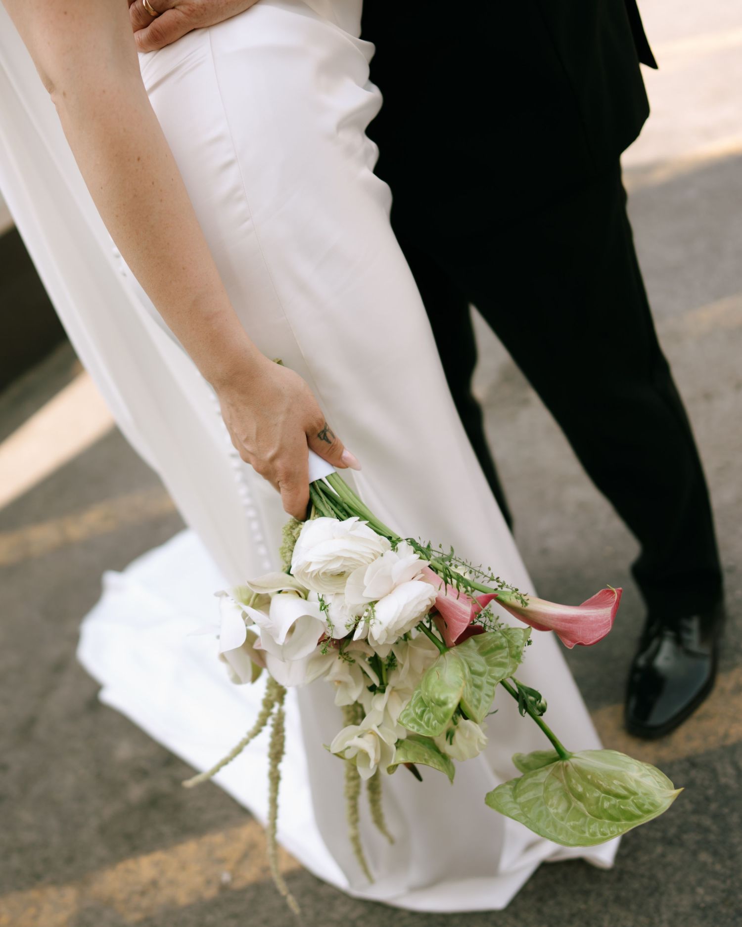 Bride in white gown holding a cascading bouquet of pink calla lilies and white roses beside groom in black suit.