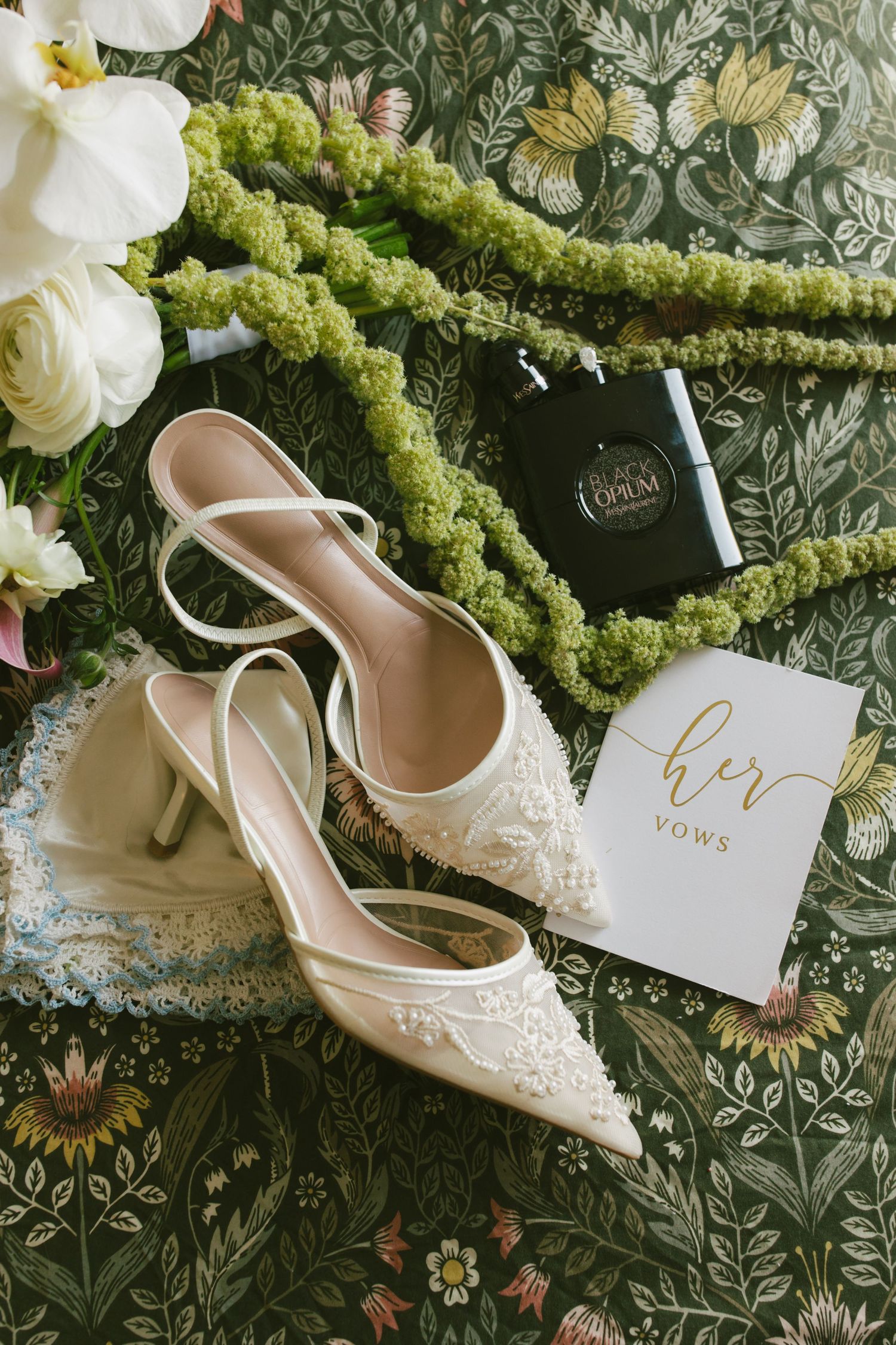 Bridal flat lay with white lace heels, YSL Black Opium perfume, her vows book, and green floral accents on floral backdrop.