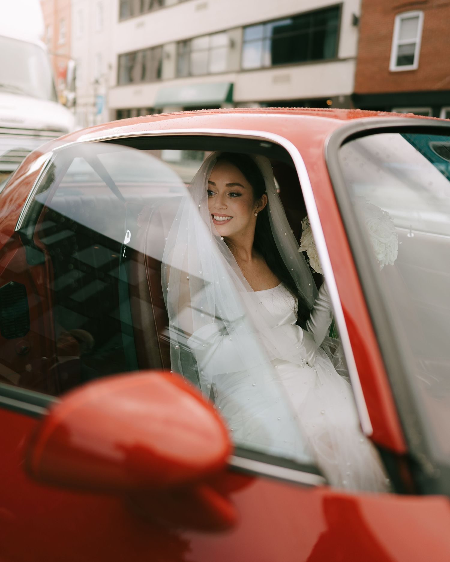 A smiling bride in a white wedding dress and veil sits inside a red car on a city street.