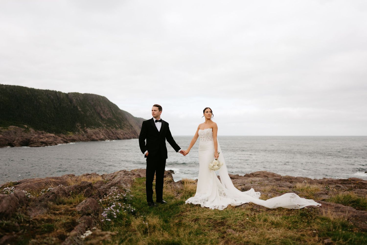 Bride and groom holding hands on rocky coastal cliffside, wearing elegant wedding attire with ocean backdrop.
