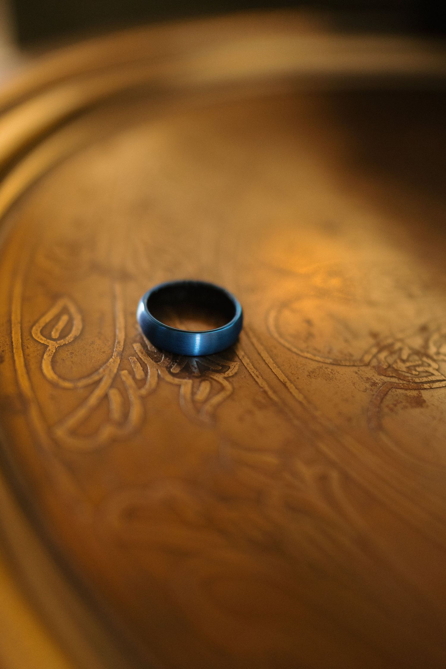 A blue tungsten ring sits on an ornate copper or brass surface with decorative engravings in warm lighting.