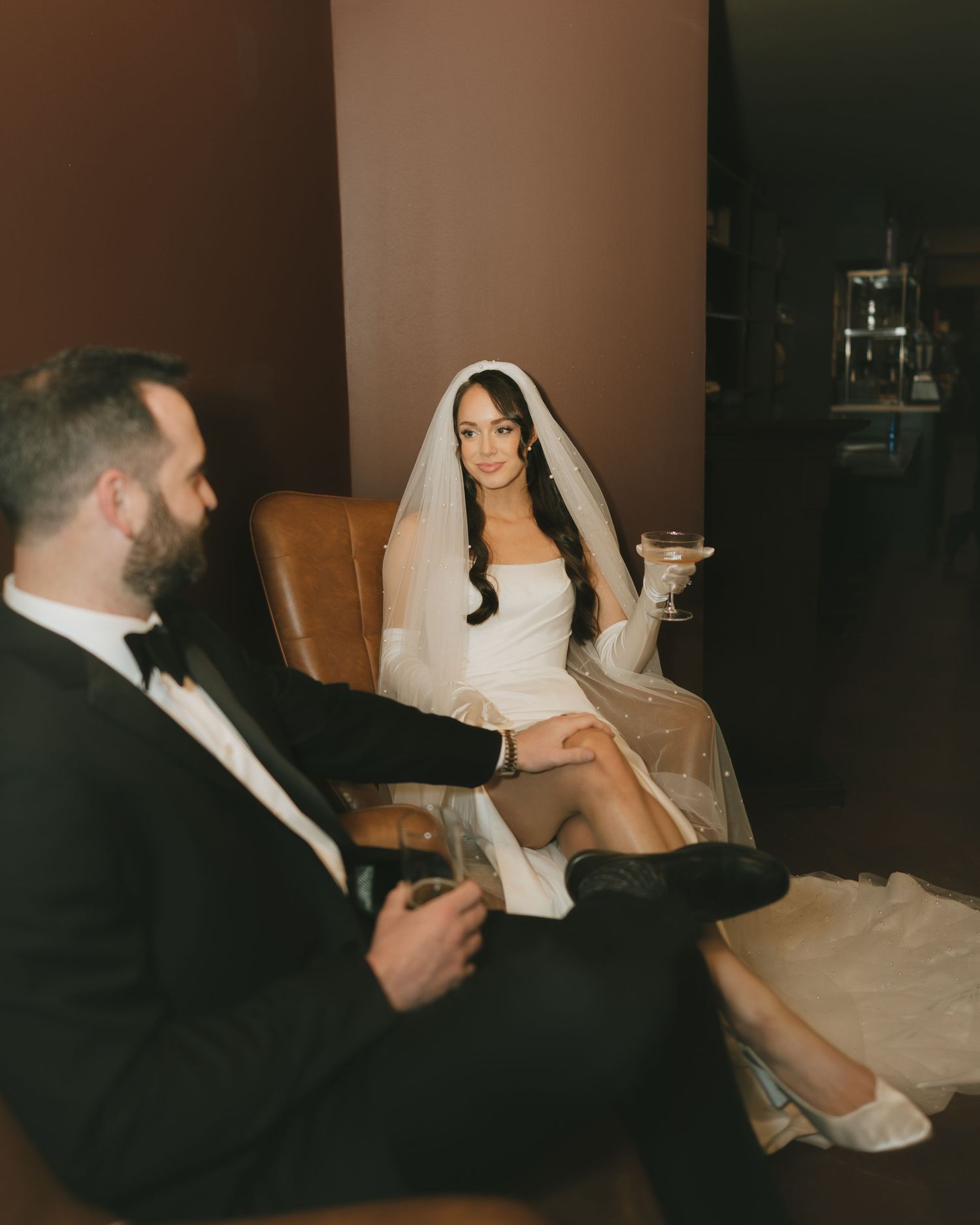 Bride in white mini dress and veil sits in leather chair holding cocktail beside groom in black tuxedo.