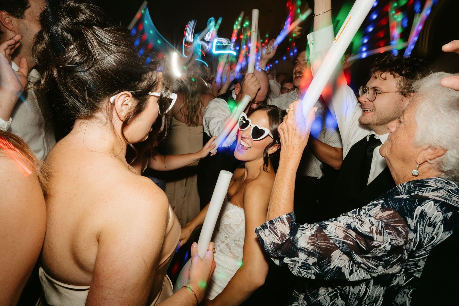 Guests dancing at a wedding reception with colorful glow sticks and LED lights in a dark venue.