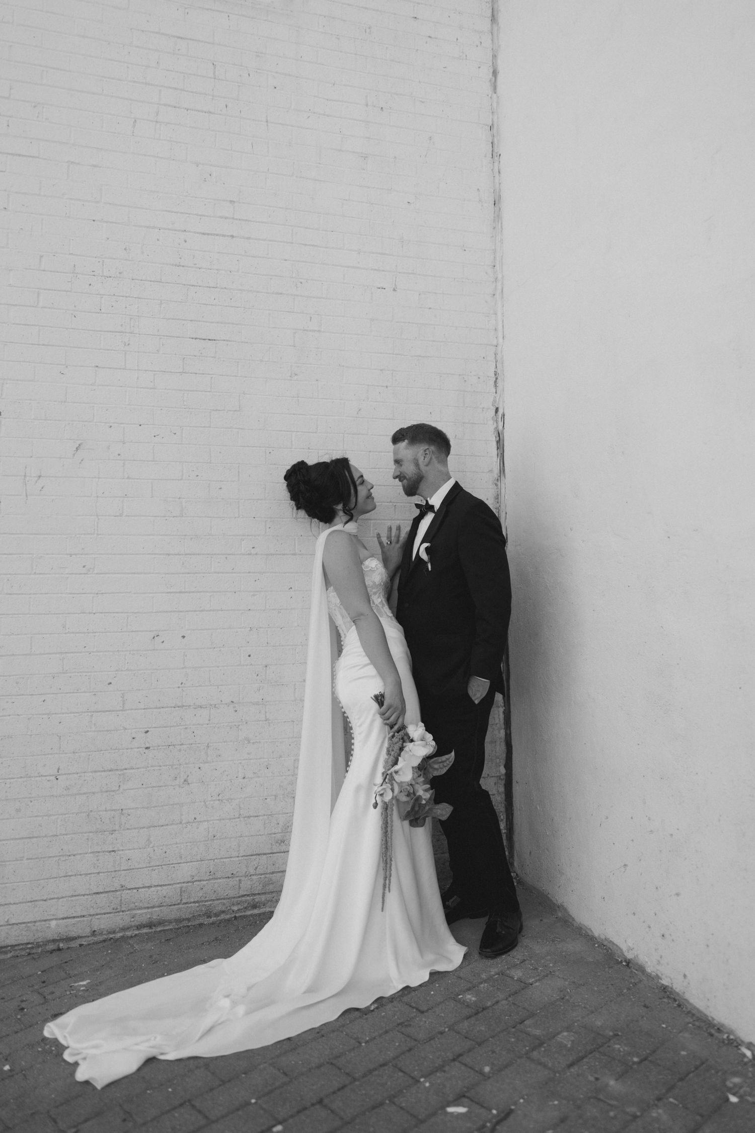 Black and white photo of a bride and groom leaning against a brick wall, gazing at each other on their wedding day.