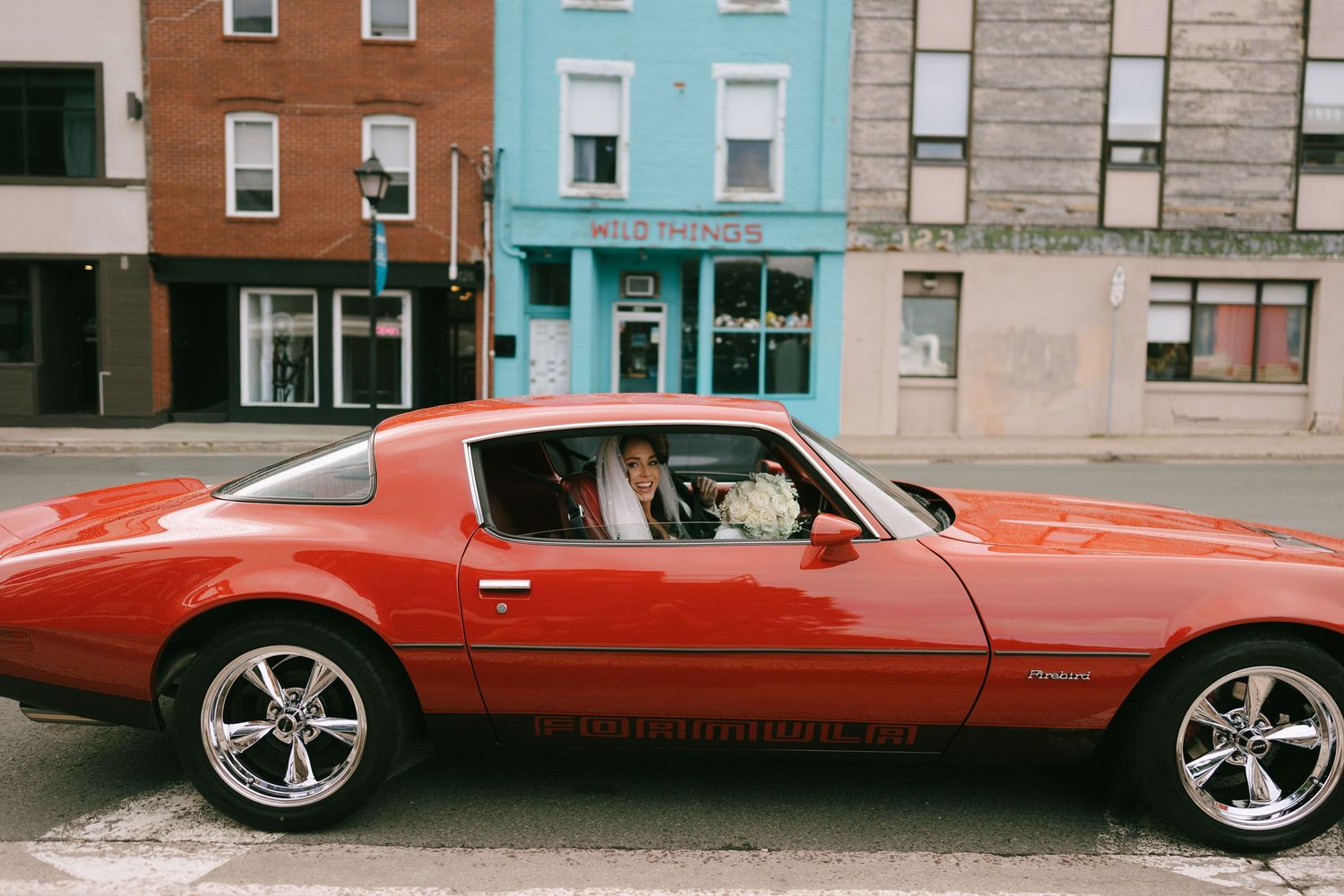 A classic red Pontiac Firebird muscle car parked on a city street with colorful buildings in the background.