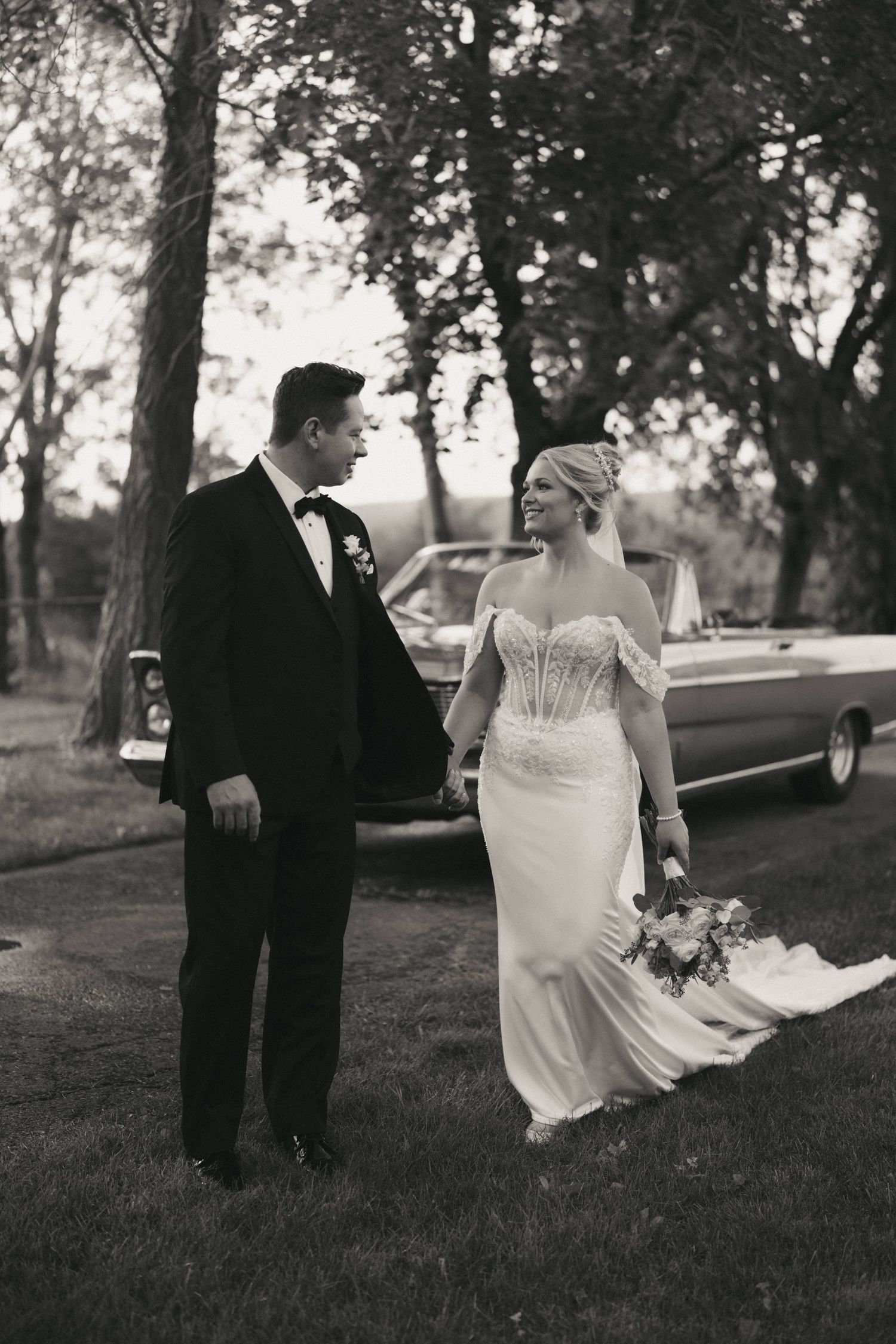 Black and white photo of bride and groom holding hands near vintage car outdoors on their wedding day.