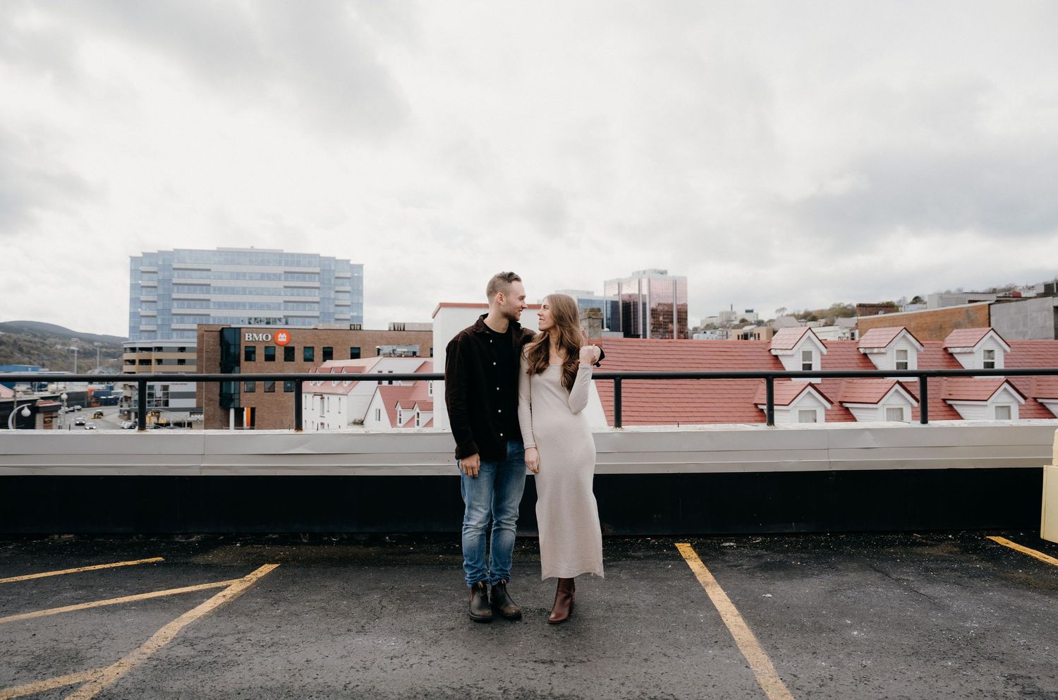 Wide angle shot of couple standing on empty parking deck with city buildings in background.