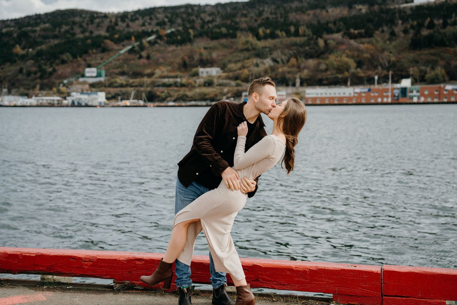 A romantic photoshoot by the waterfront with a bridge and mountainous backdrop on an overcast day.