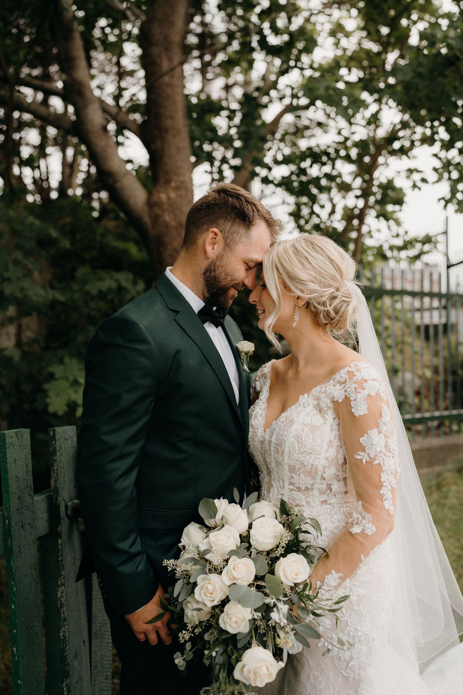 A couple sharing an intimate moment surrounded by trees with a white floral bouquet.
