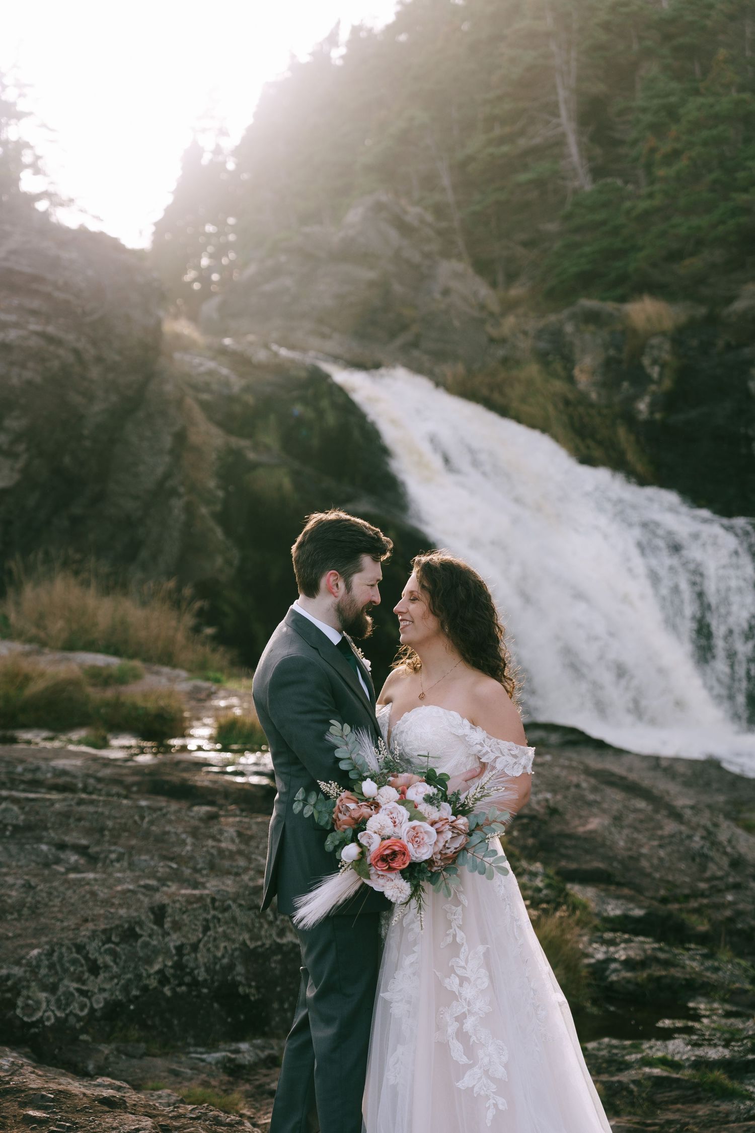 Romantic wedding couple embracing by dramatic waterfall in forest setting.