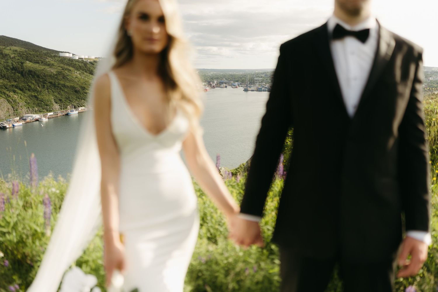 Couple in wedding attire poses in front of scenic coastal landscape with blooming wildflowers.