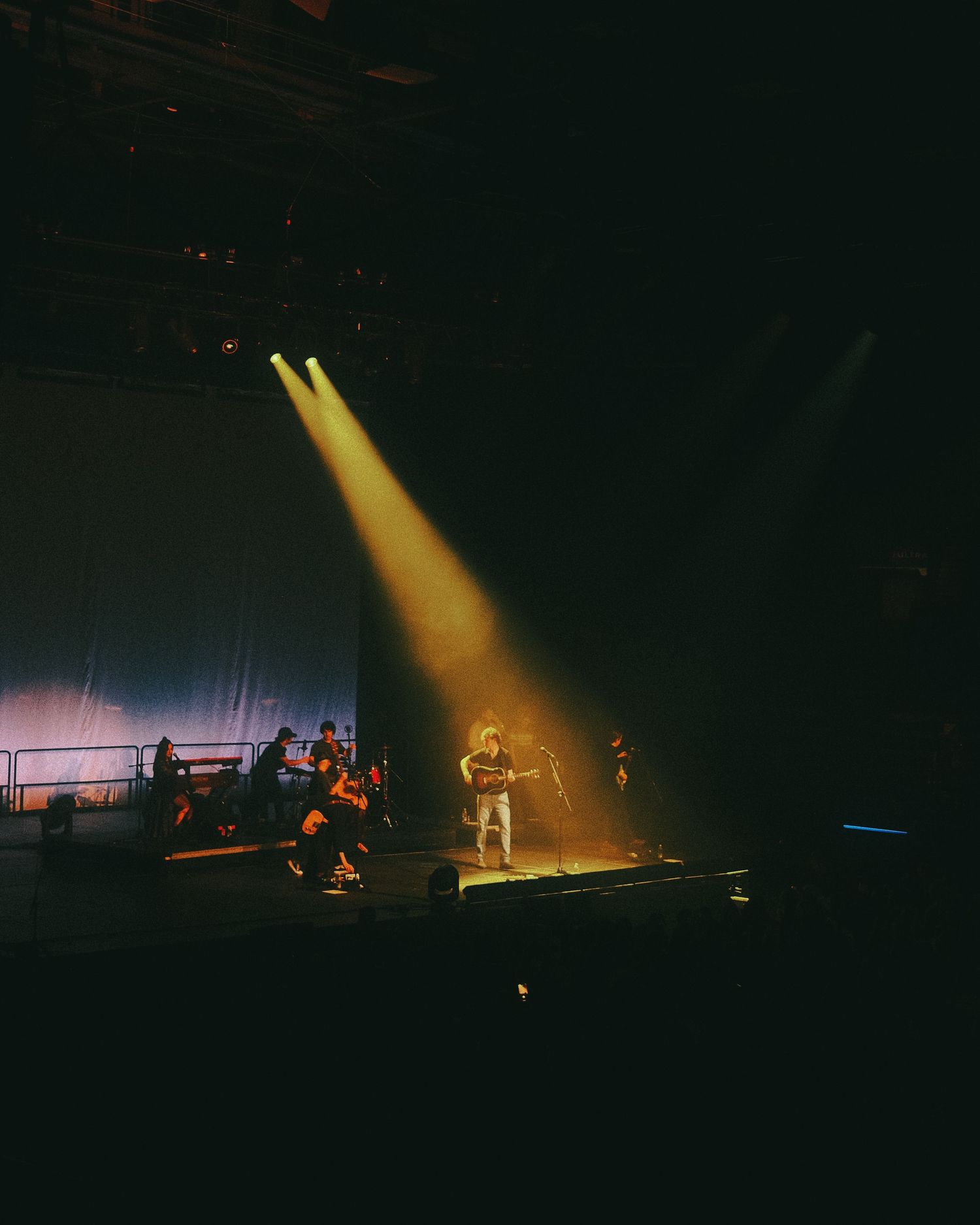 Dramatic concert stage lighting illuminates performers through yellow spotlights in a dark venue during a live show.