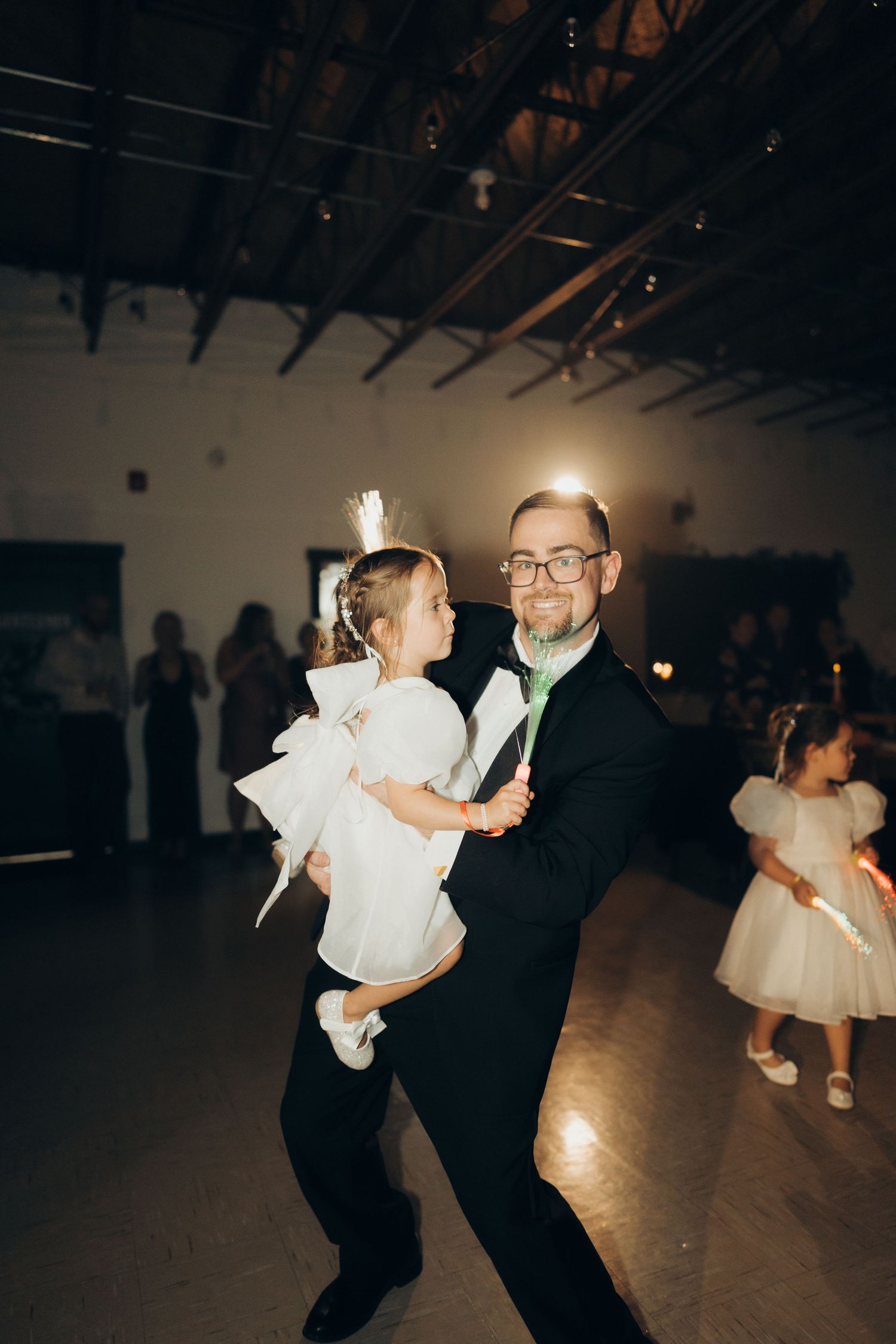 A father and daughter dance together at a formal event in a dimly lit venue with white dresses visible in background.