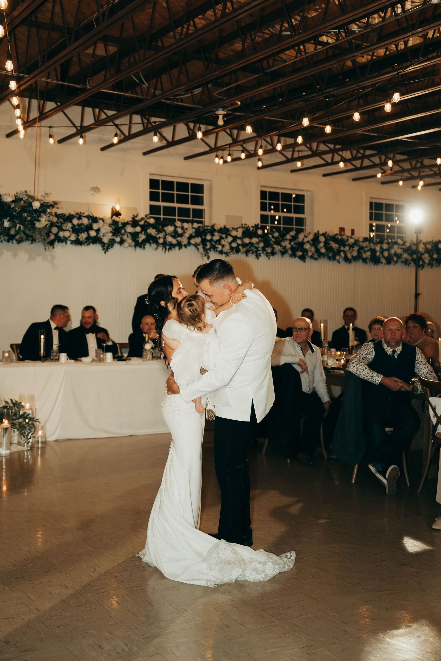 A couple shares their first dance at a wedding reception in an industrial-style venue with string lights overhead.