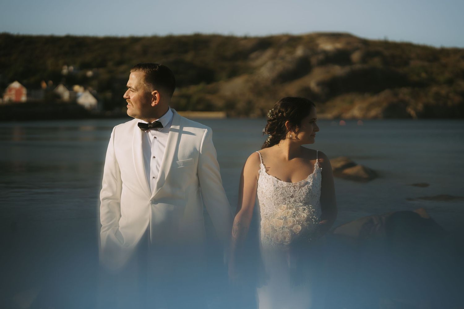 A couple in wedding attire stands together on a rocky shoreline at sunset with a serene lake in the background.