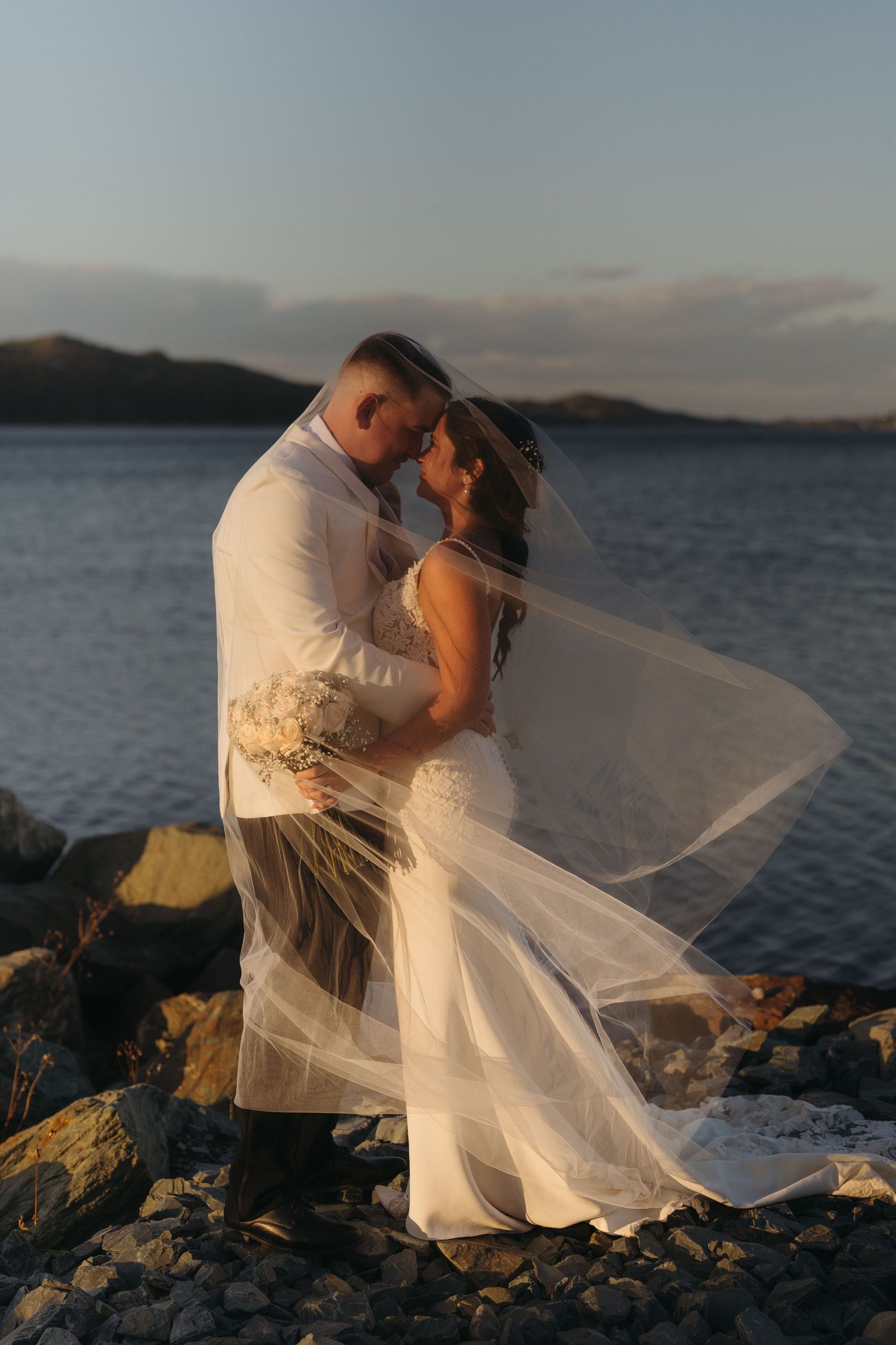 A romantic wedding couple embraces on rocky coastal cliffs at sunset as the bride's veil flows dramatically in the wind.