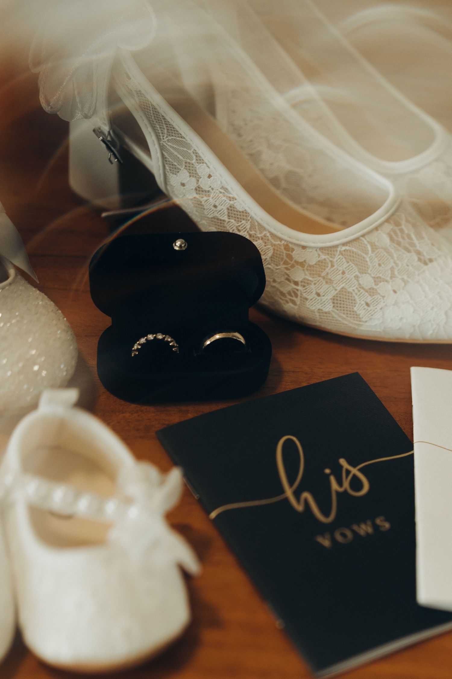 Wedding vows books in black with gold lettering sit beside delicate white lace bridal shoes on wooden surface.