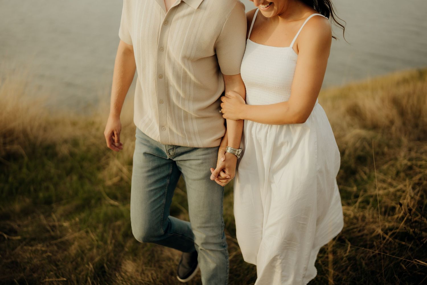 A couple holds hands while walking through a golden field at sunset wearing casual engagement photo attire.