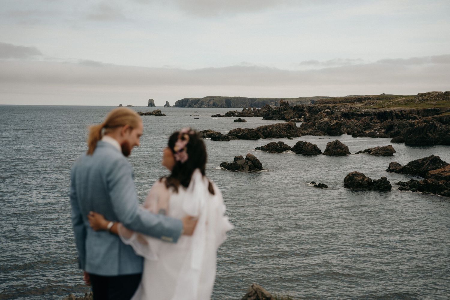 A romantic couple embraces on a rocky coastline with dramatic ocean views and rugged cliffs in the background.