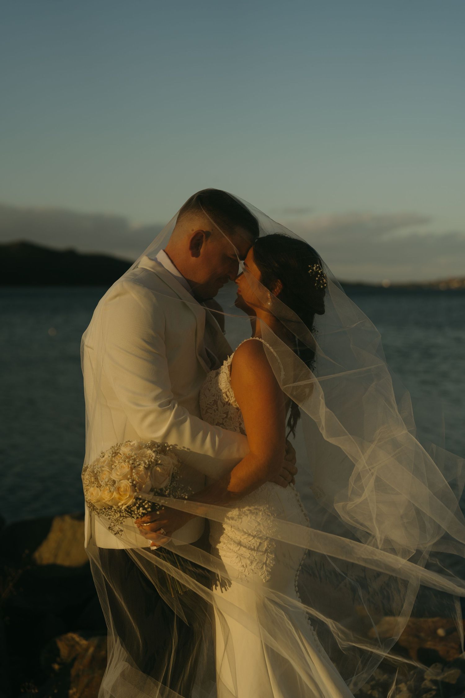 A romantic beach wedding couple embraces as the bride's veil flows in the wind during golden hour at sunset.