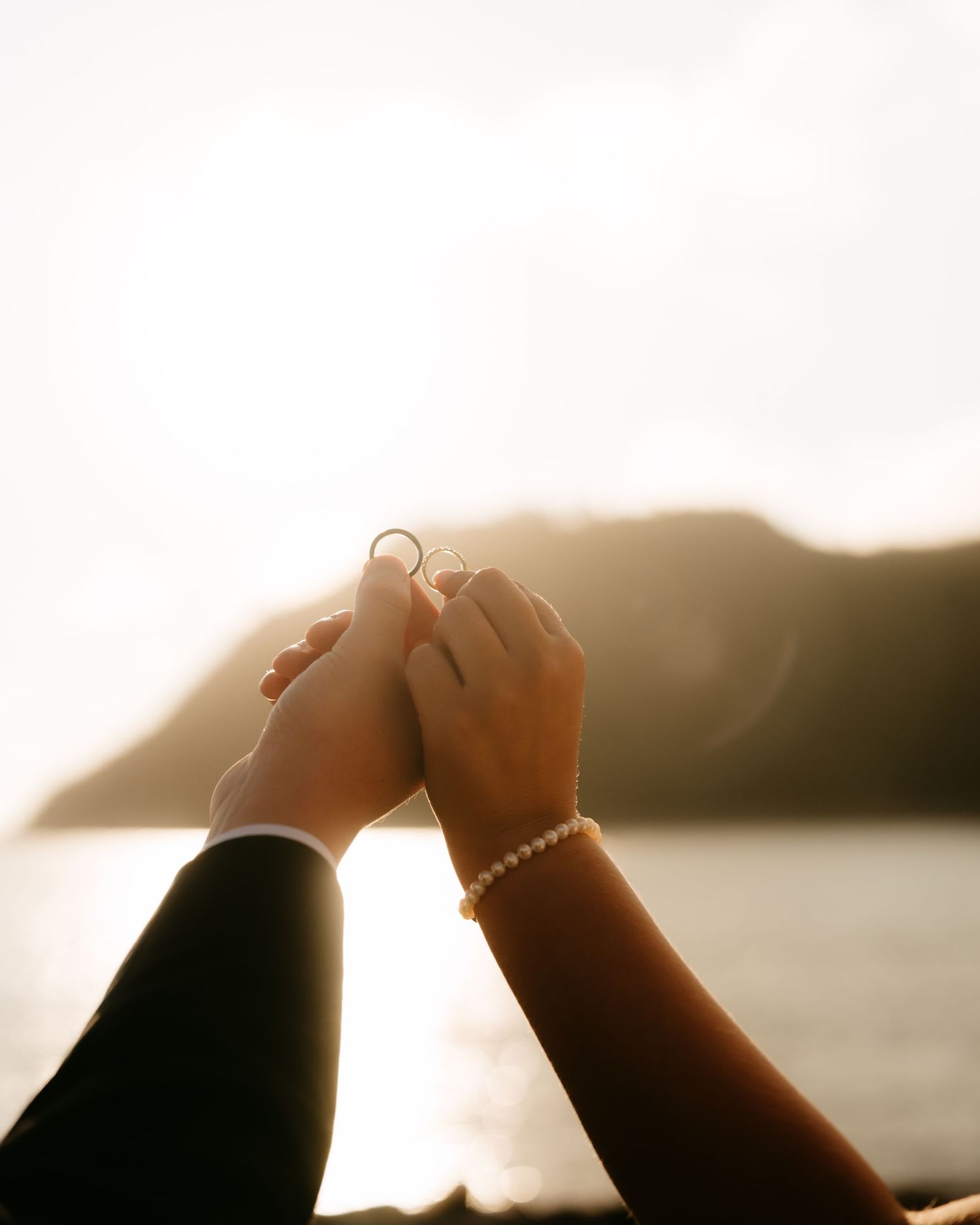 Couple holding hands with wedding rings at golden hour by the ocean with a mountain backdrop.