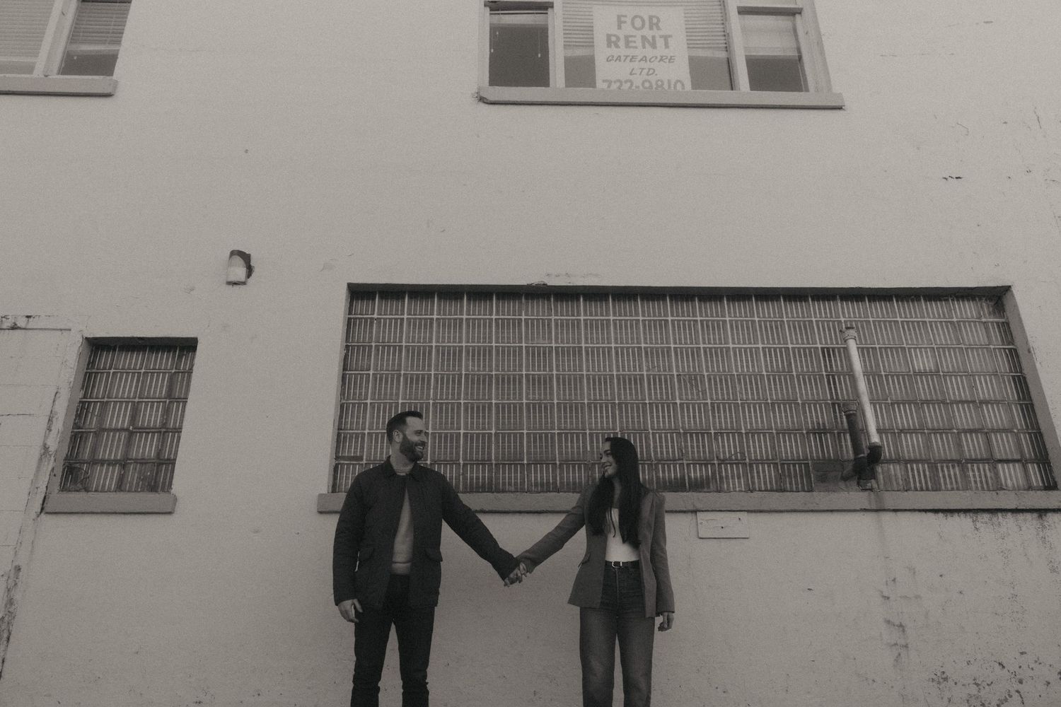 Black and white photo of a couple holding hands in front of a large industrial building with a for rent sign.