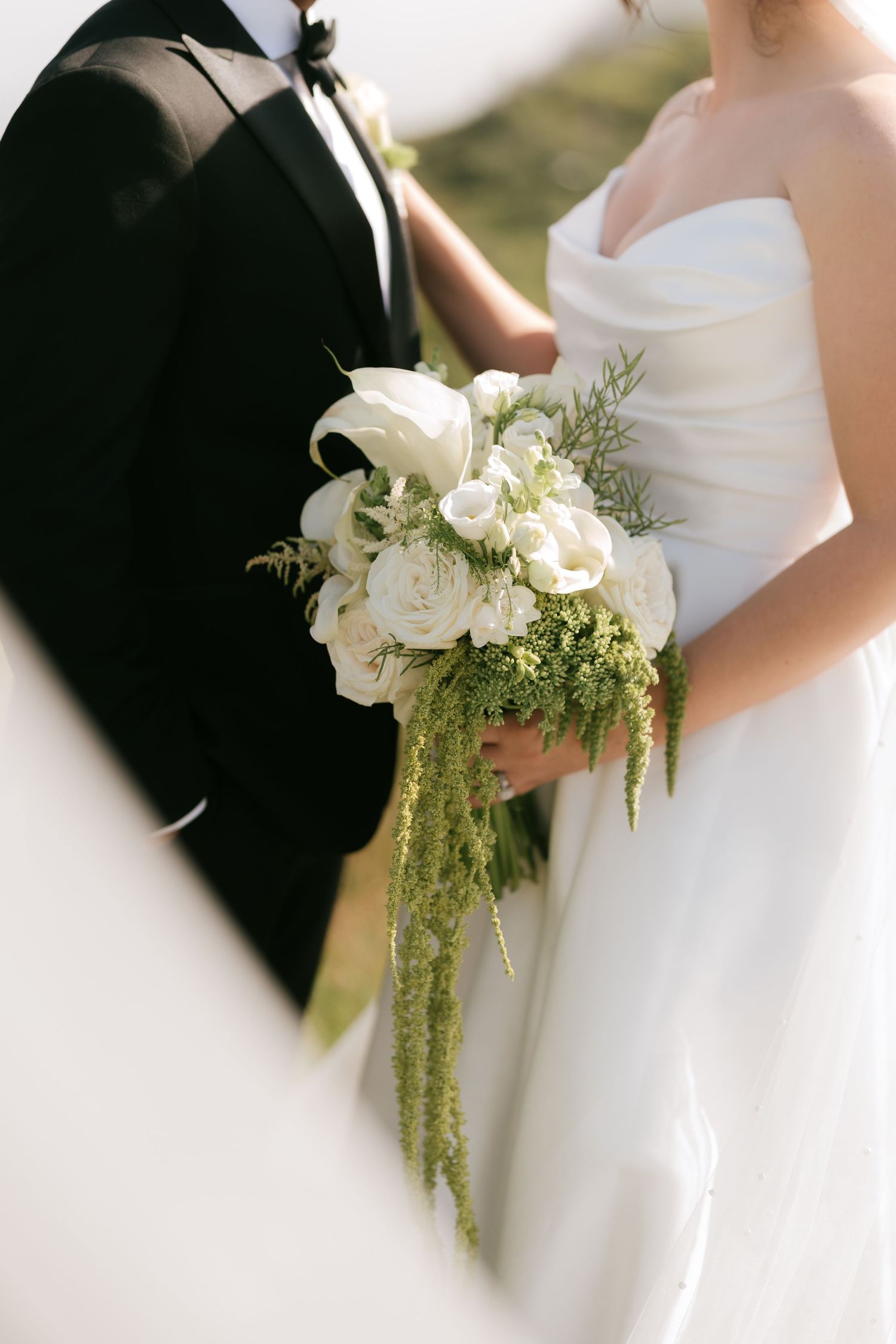 Bride holding white rose and greenery wedding bouquet with cascading amaranthus beside groom in black tuxedo.