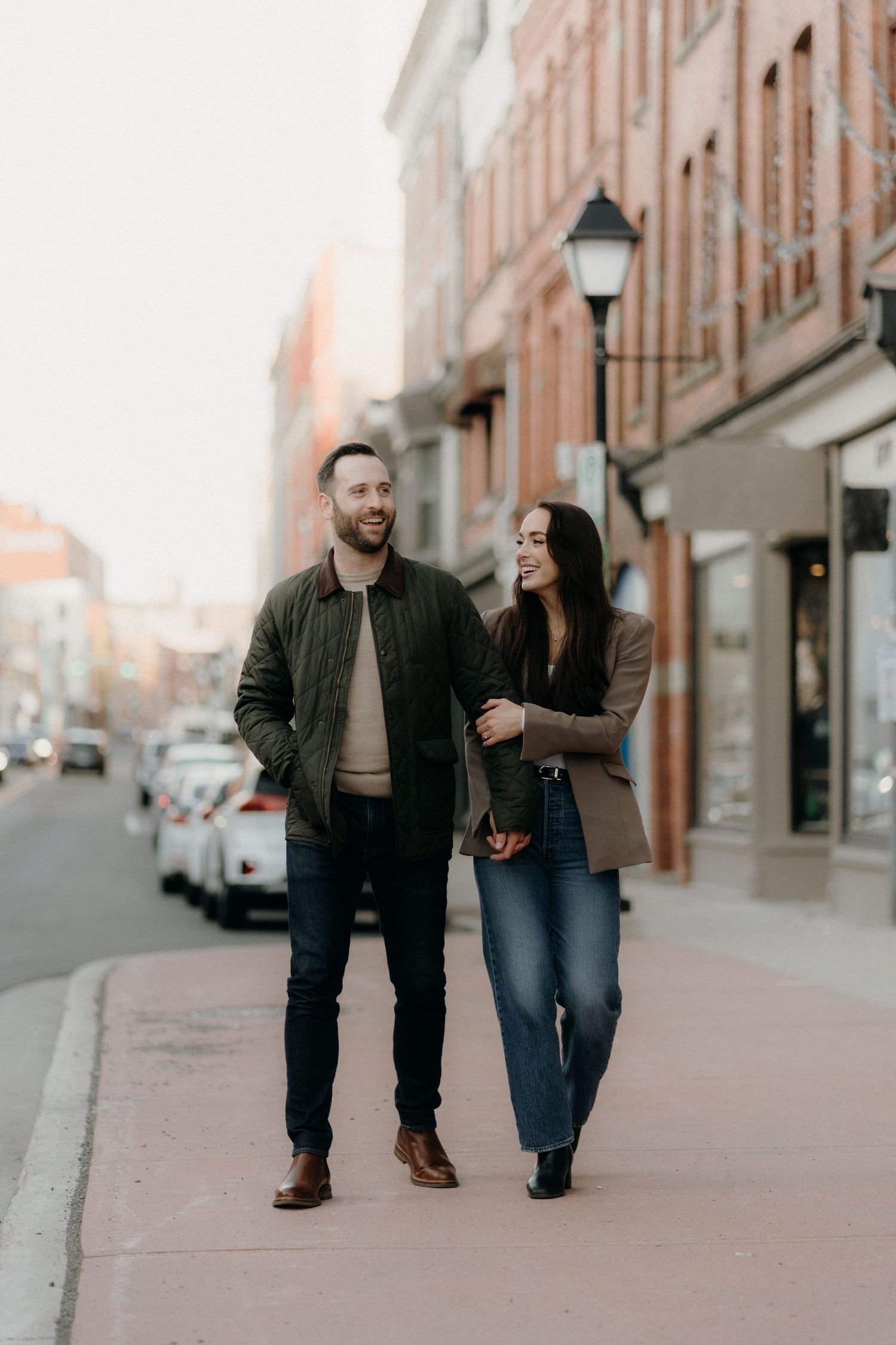 A couple walks arm-in-arm along a charming urban sidewalk, smiling at each other in casual autumn outfits.