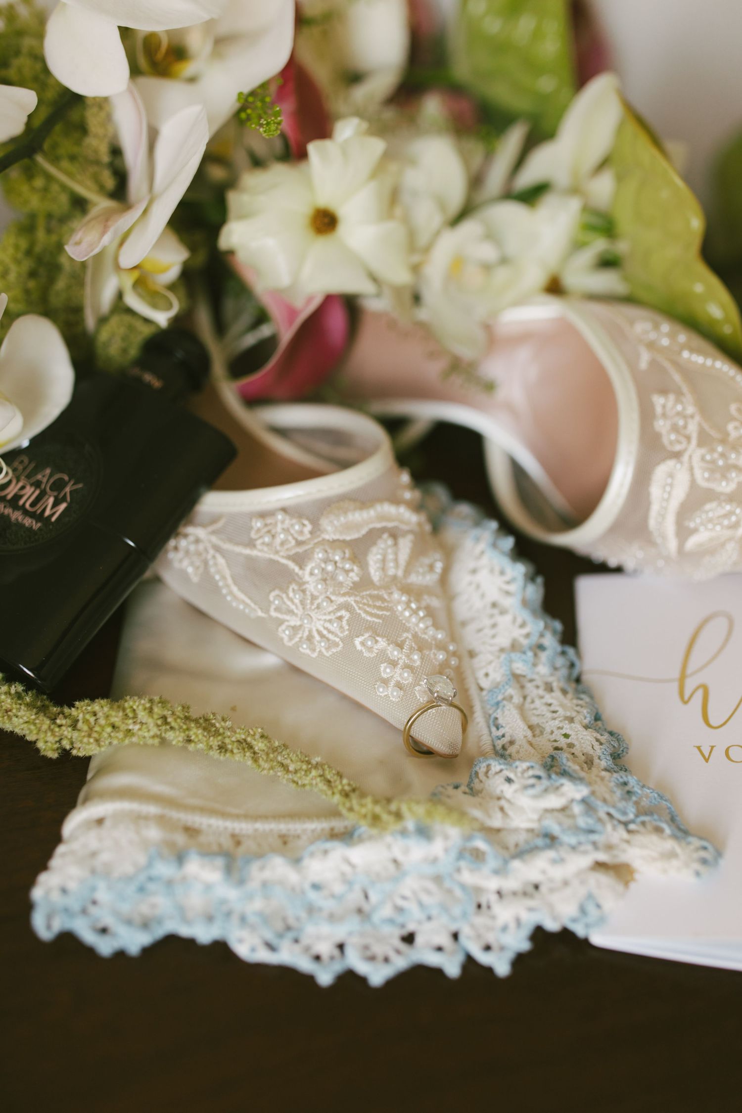 Bridal flat lay with lace garter, gold ring, embroidered shoes, white flowers, and vow booklet on dark surface.