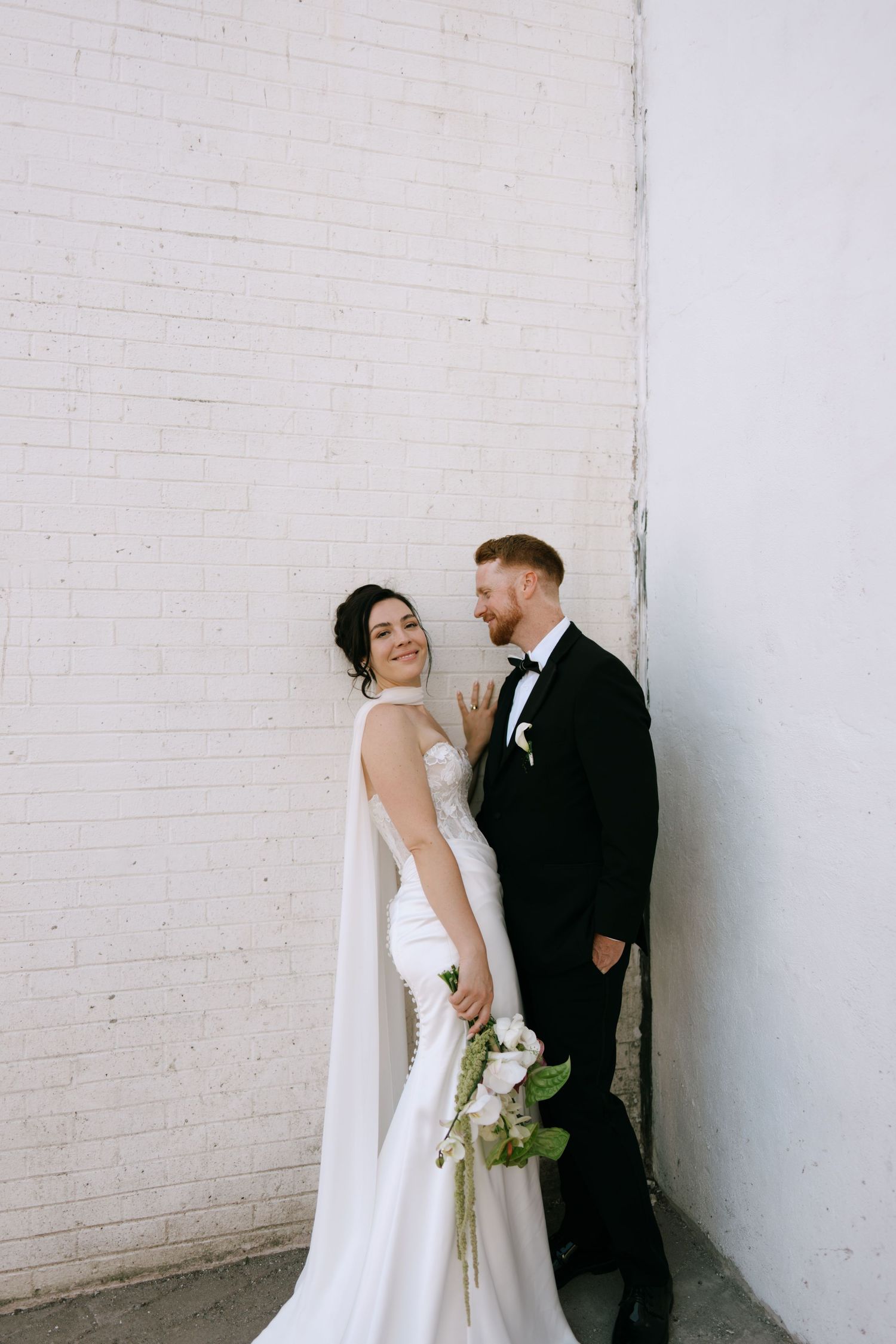 Bride in white gown holds bouquet while groom in black tuxedo stands against white brick wall.