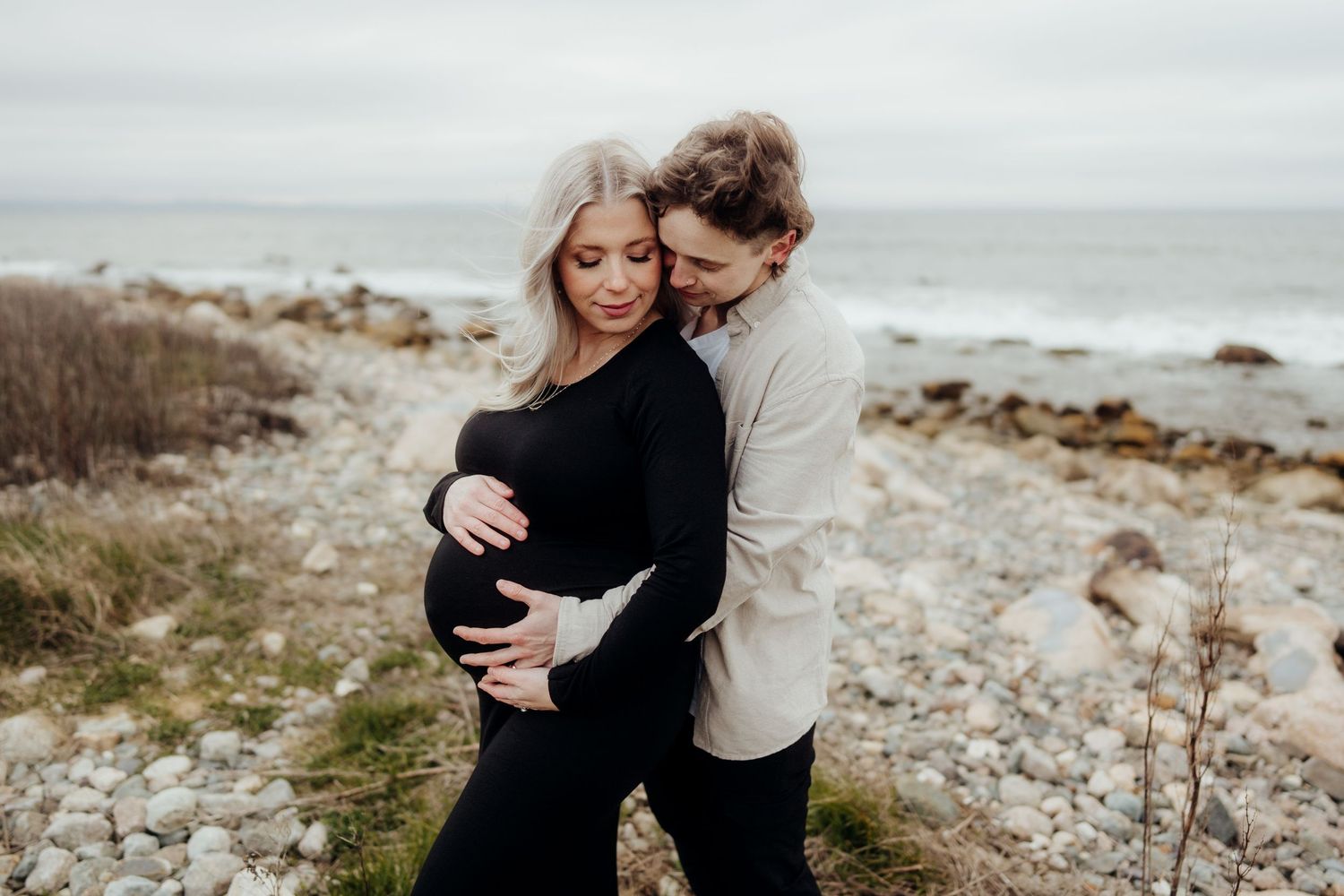 Romantic coastal maternity photos with couple embracing on rocky beach.