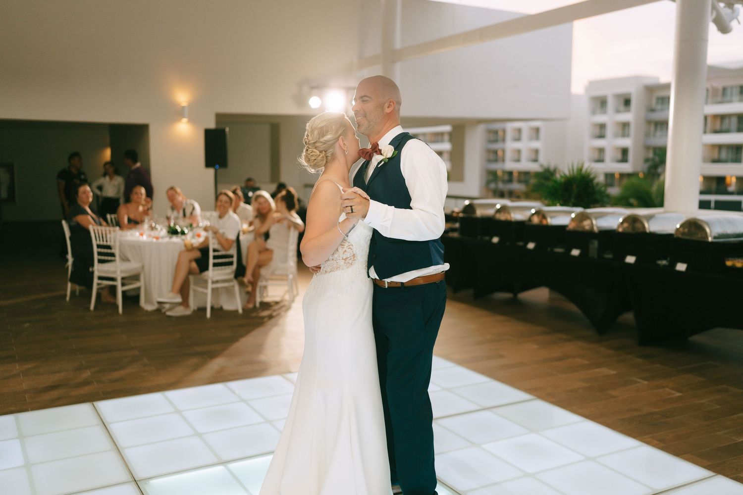 Wedding couple dancing in an elegant white ballroom with tables and decorations.