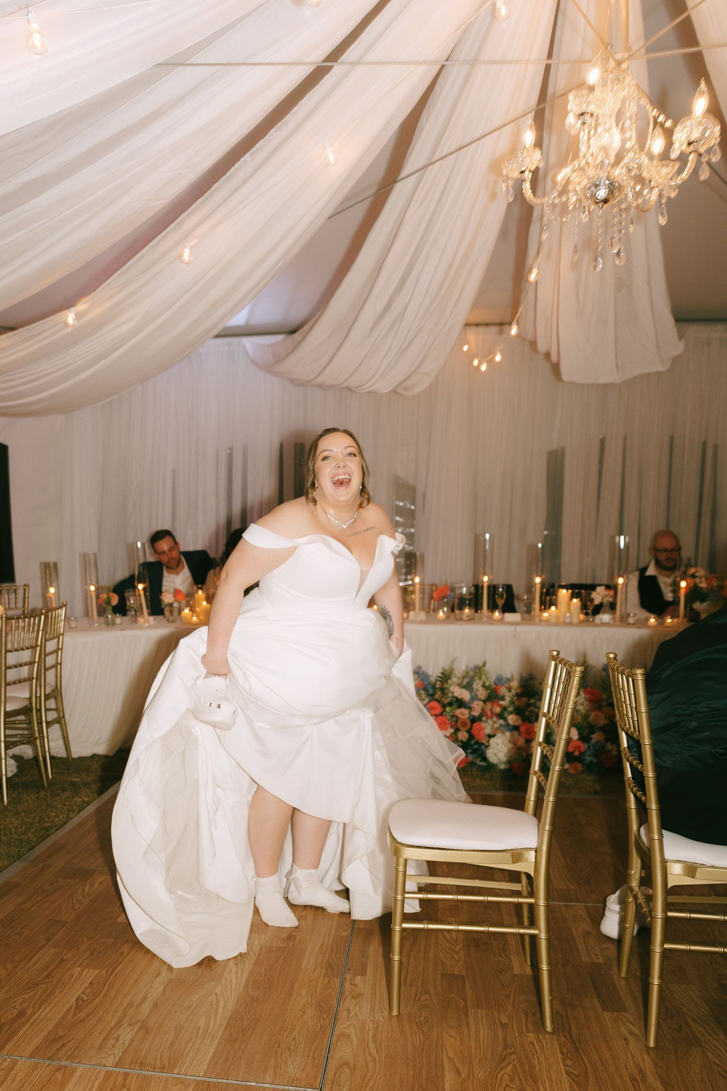 A wedding reception venue decorated with white draping, chandeliers, and gold chairs.