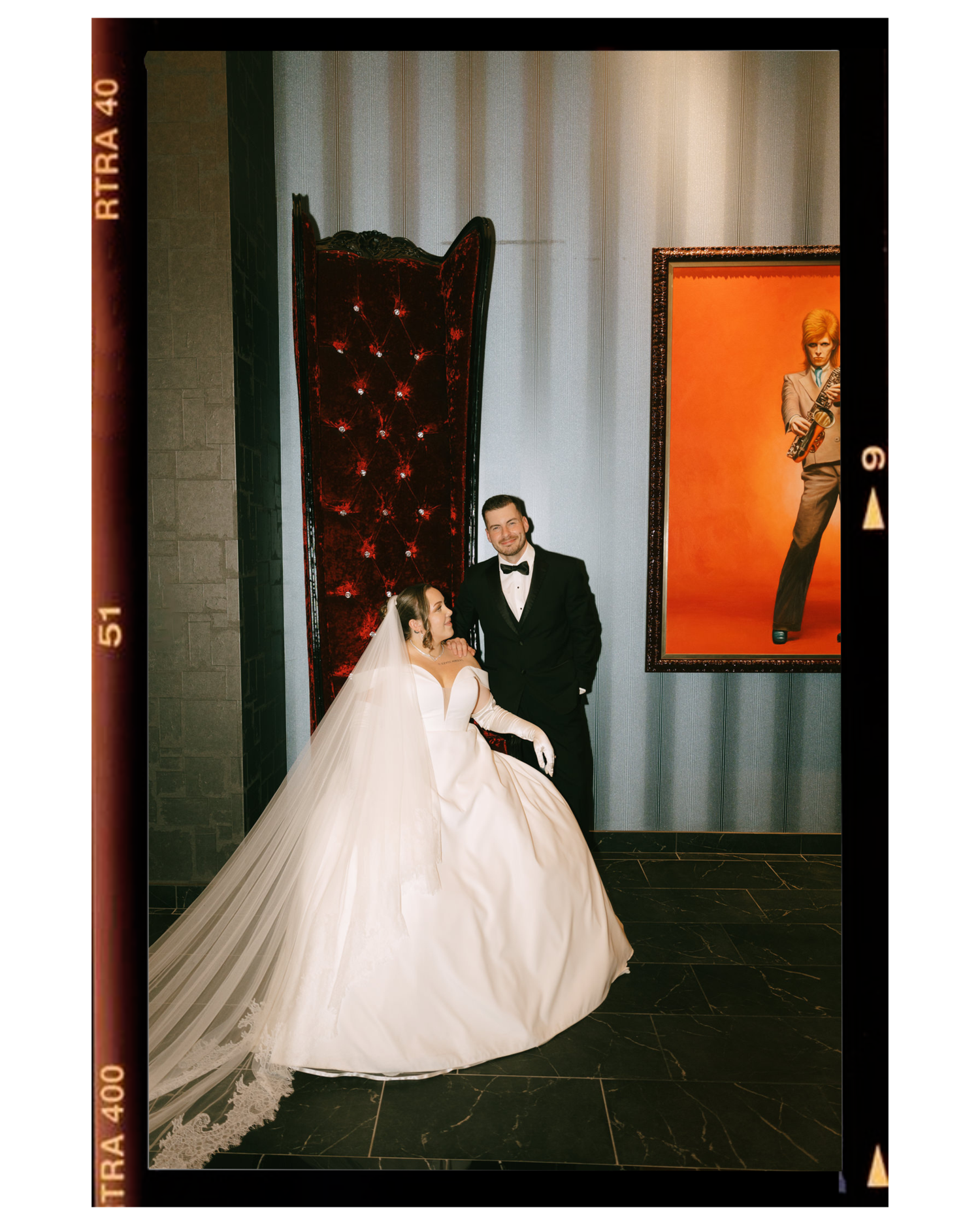 Vintage-style film photo of a wedding couple posing against an ornate wall with orange accents.