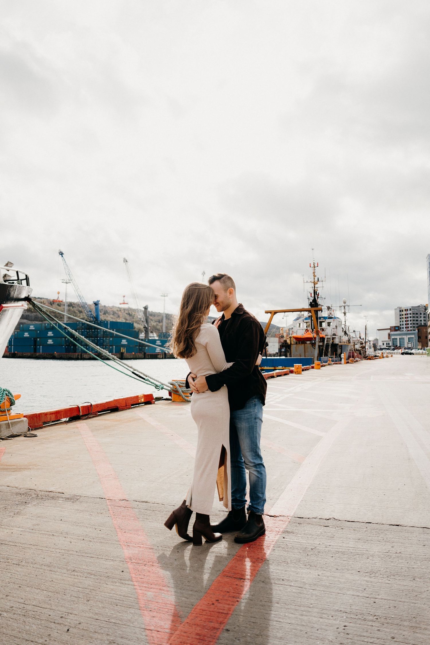 Couple dancing on waterfront pier with large vessels and industrial cranes in background.