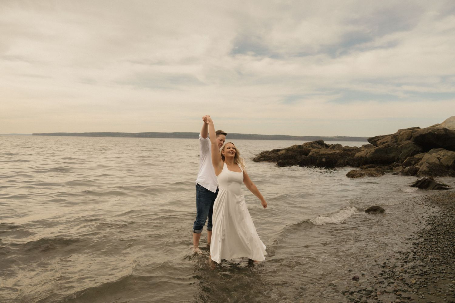 Romantic couple in formal wear embracing on a rocky shoreline during golden hour with waves crashing behind them.