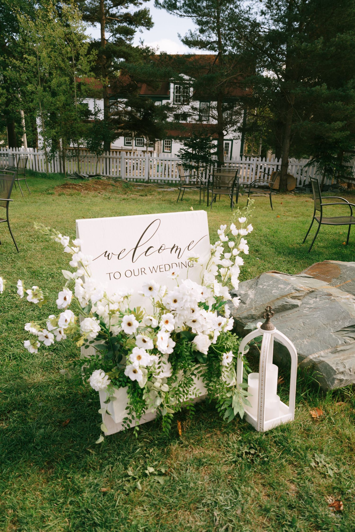 A series of white wedding welcome signs with floral arrangements displayed on rocks in an outdoor garden setting.