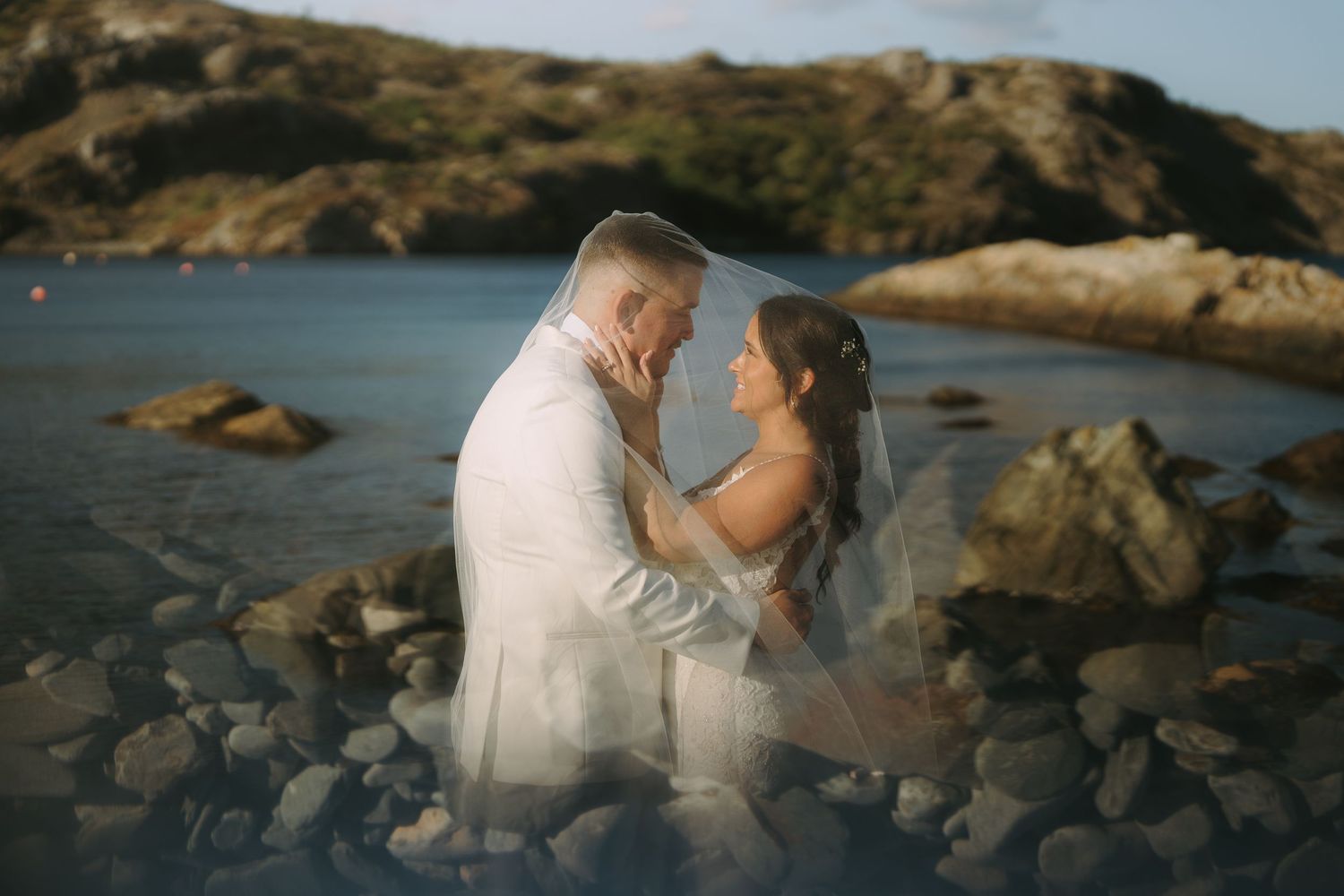 A romantic couple in white attire shares tender moments along a rocky coastal shoreline at sunset.
