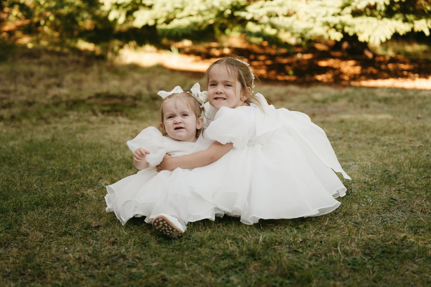 A child in a flowing white dress lies playfully on green grass during a sunny outdoor photo session.