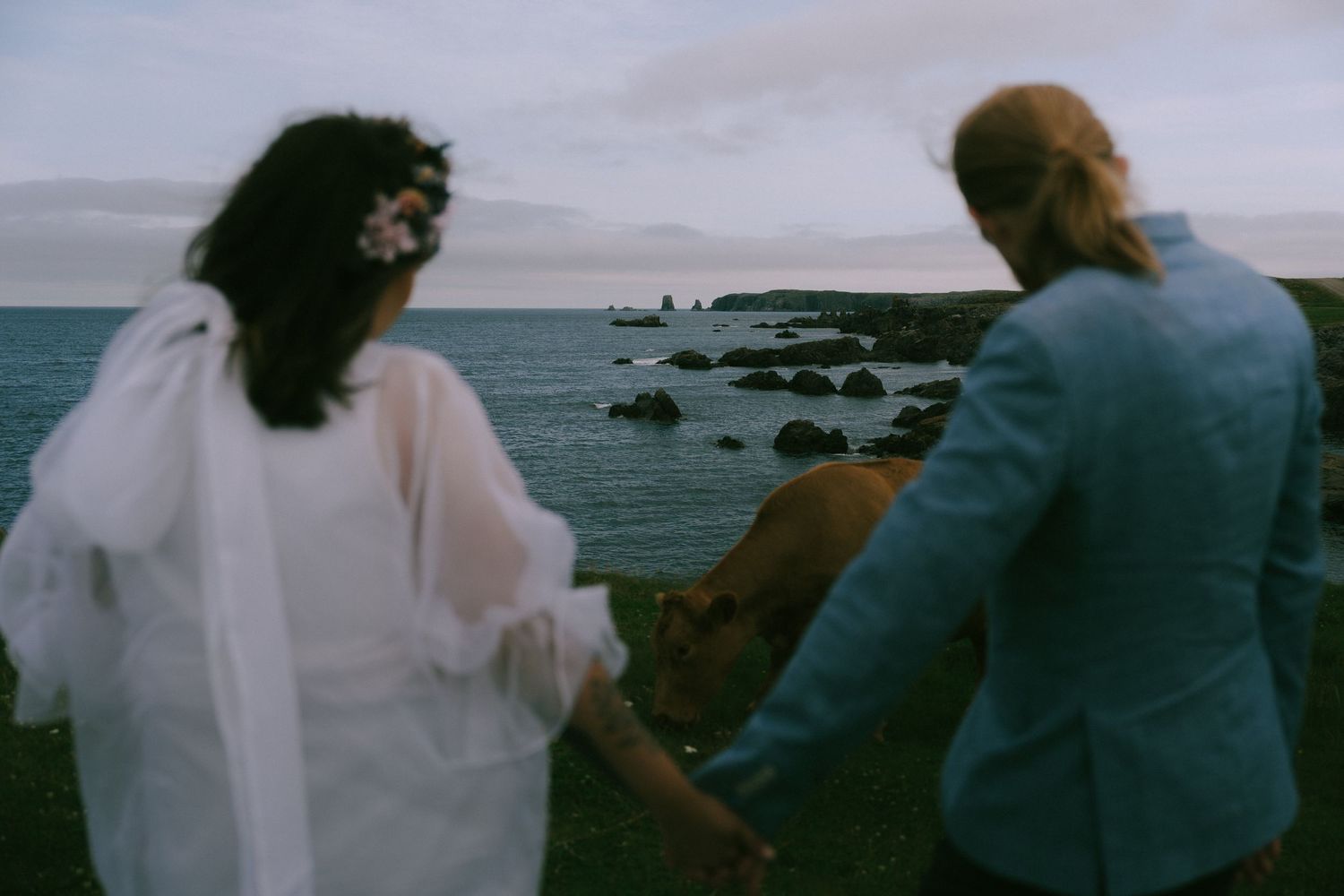 Two people in wedding attire stand at a coastal overlook watching boats on the ocean horizon.