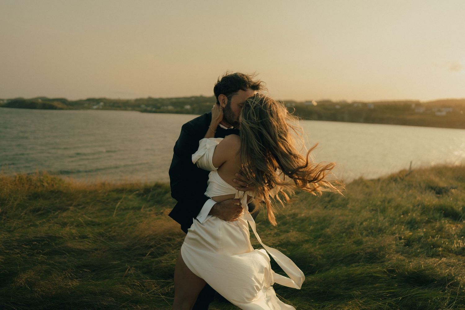 A couple shares an intimate moment at sunset on a grassy hilltop overlooking a scenic landscape.