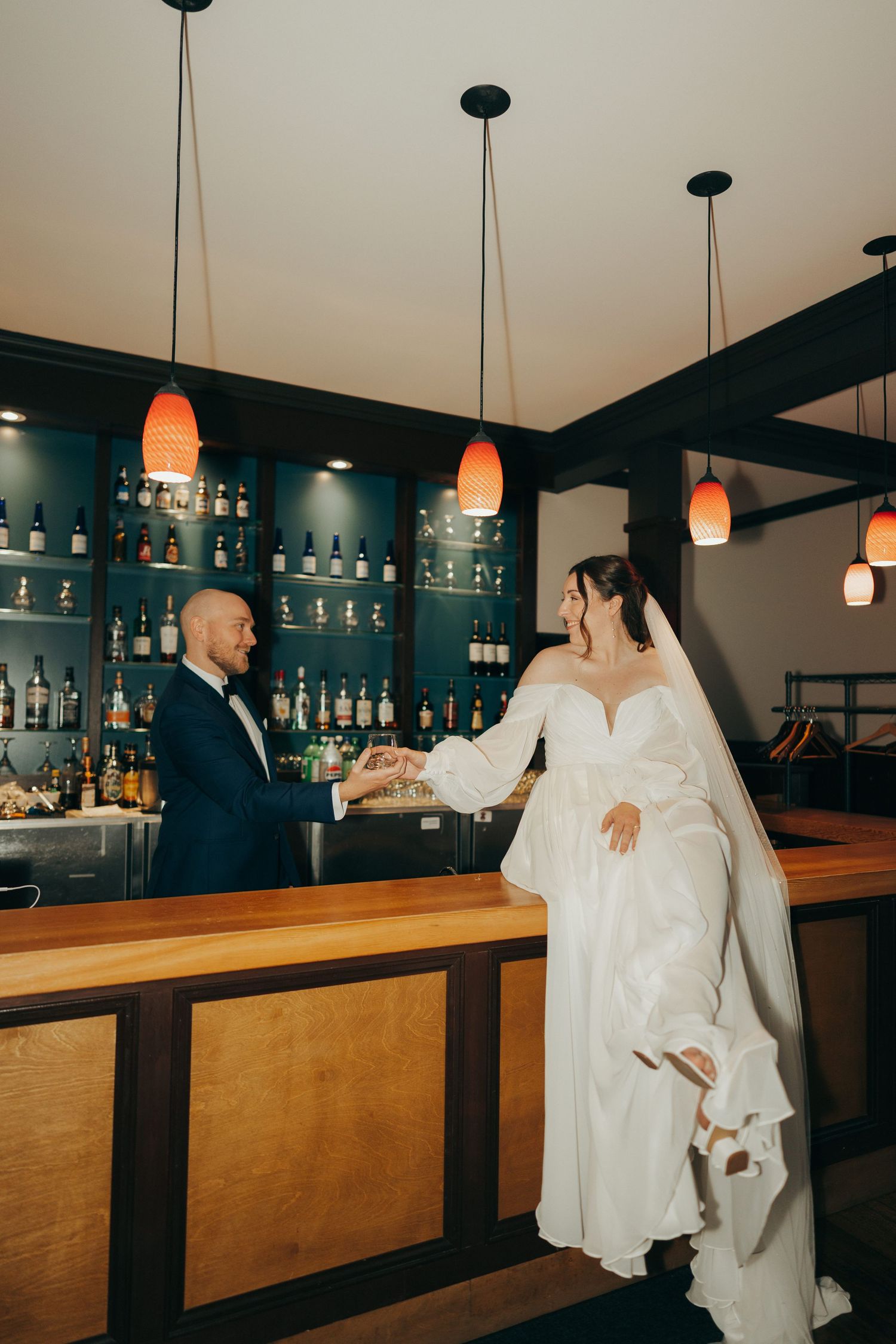 A couple in wedding attire shares a romantic moment at a modern bar with orange pendant lights and wooden countertops.