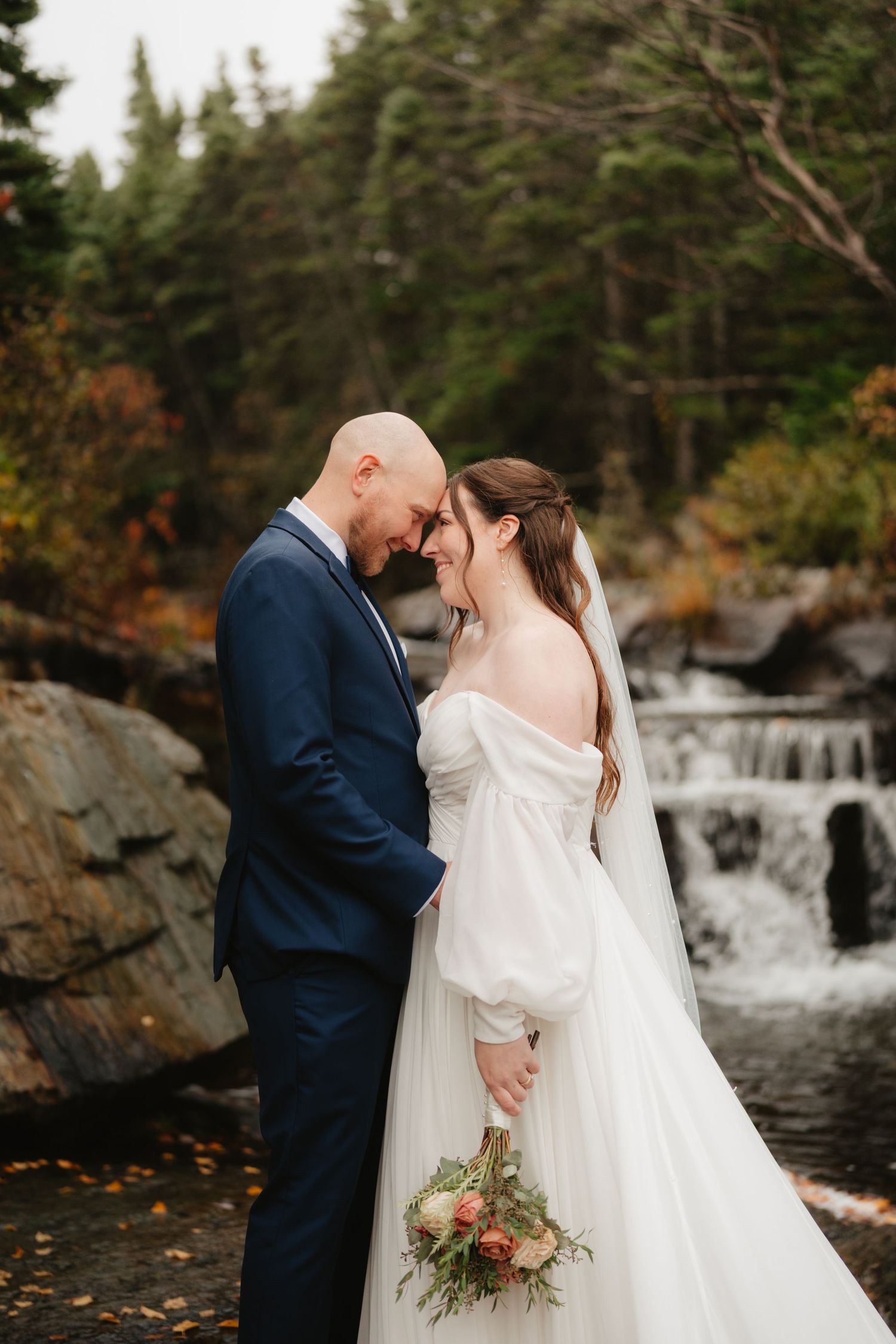 A romantic wedding couple embraces by a cascading waterfall surrounded by autumn foliage in a natural setting.