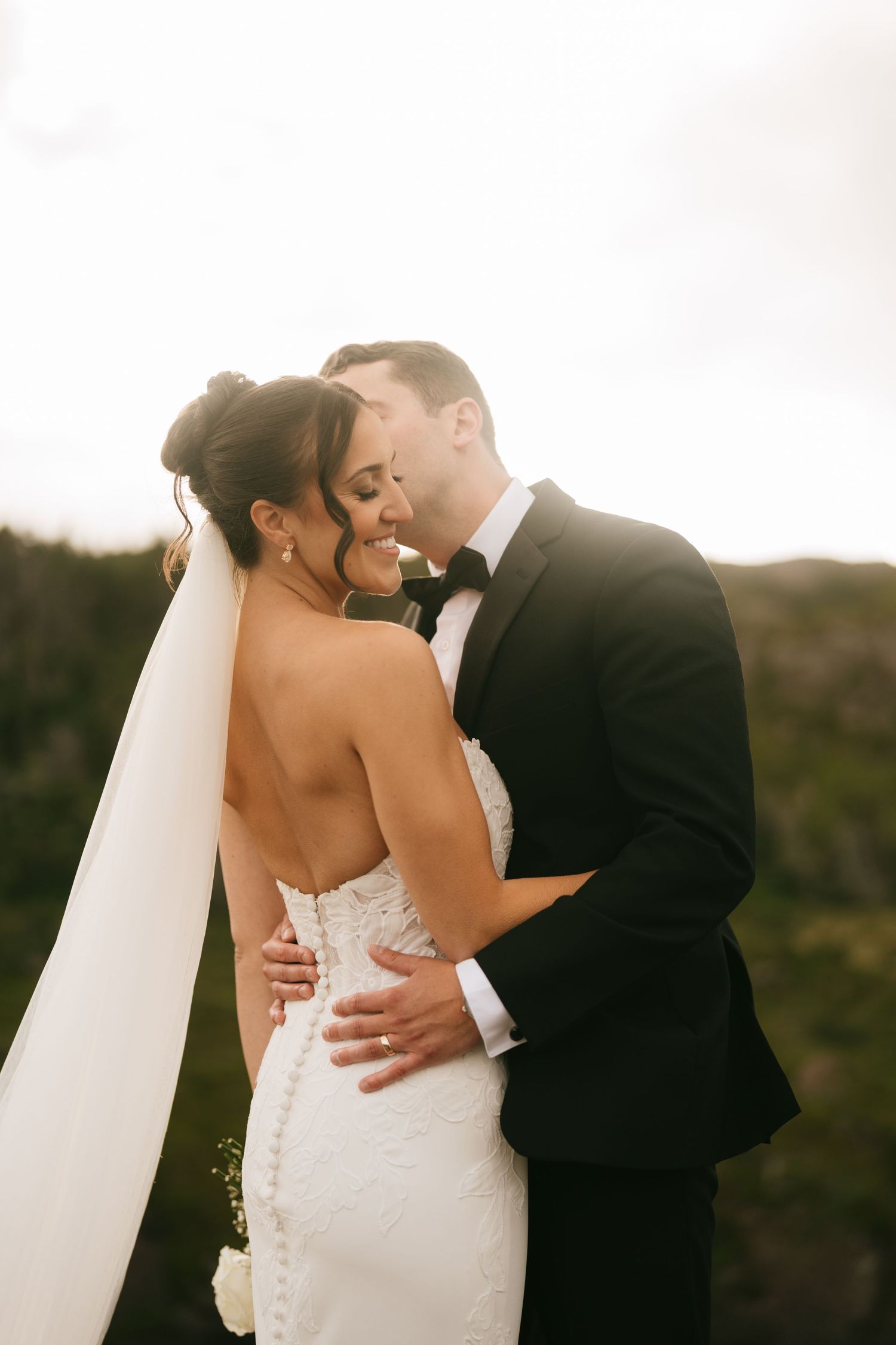 A bride and groom share an intimate embrace outdoors, with the bride in a lace gown and veil, groom in a black tuxedo.
