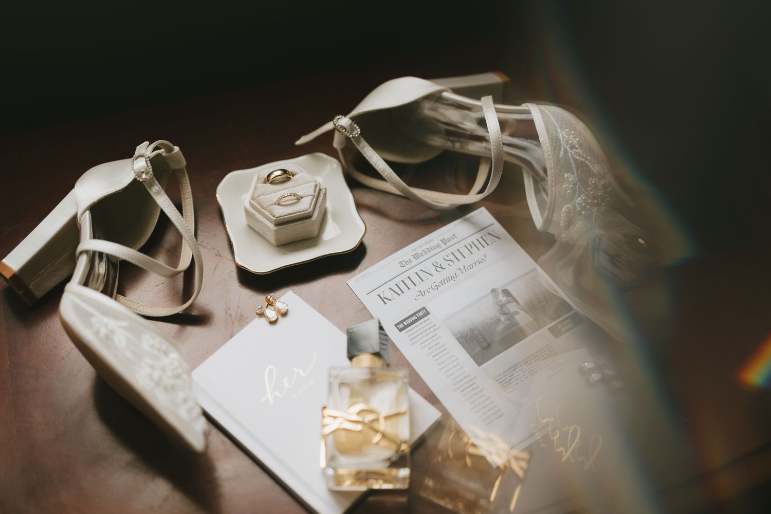 Elegant wedding flat lay with bridal heels, ring box, invitations, perfume, and gold accessories on dark surface.