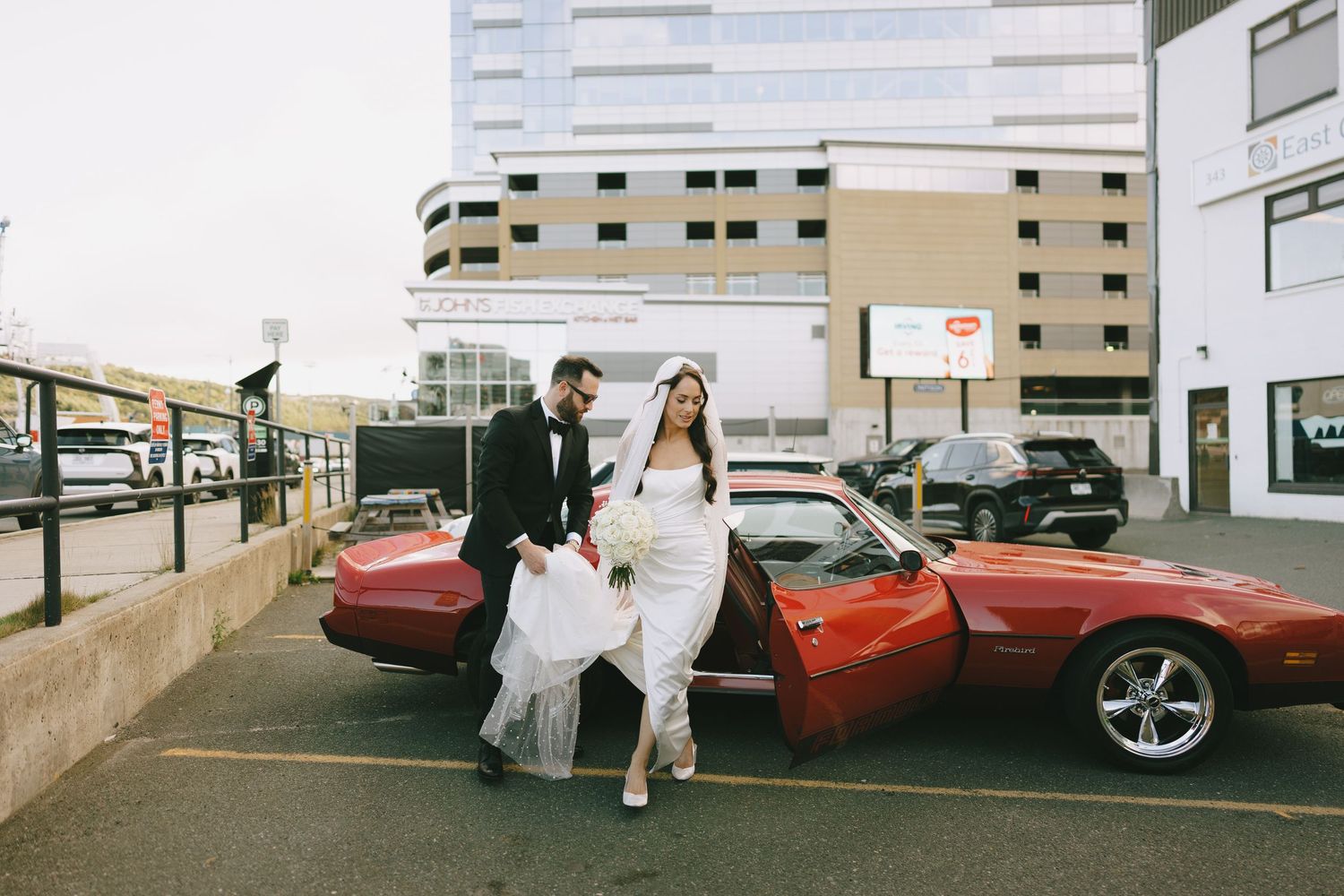 Bride and groom exit a classic red Corvette convertible in a parking lot, with the bride holding her white wedding dress.