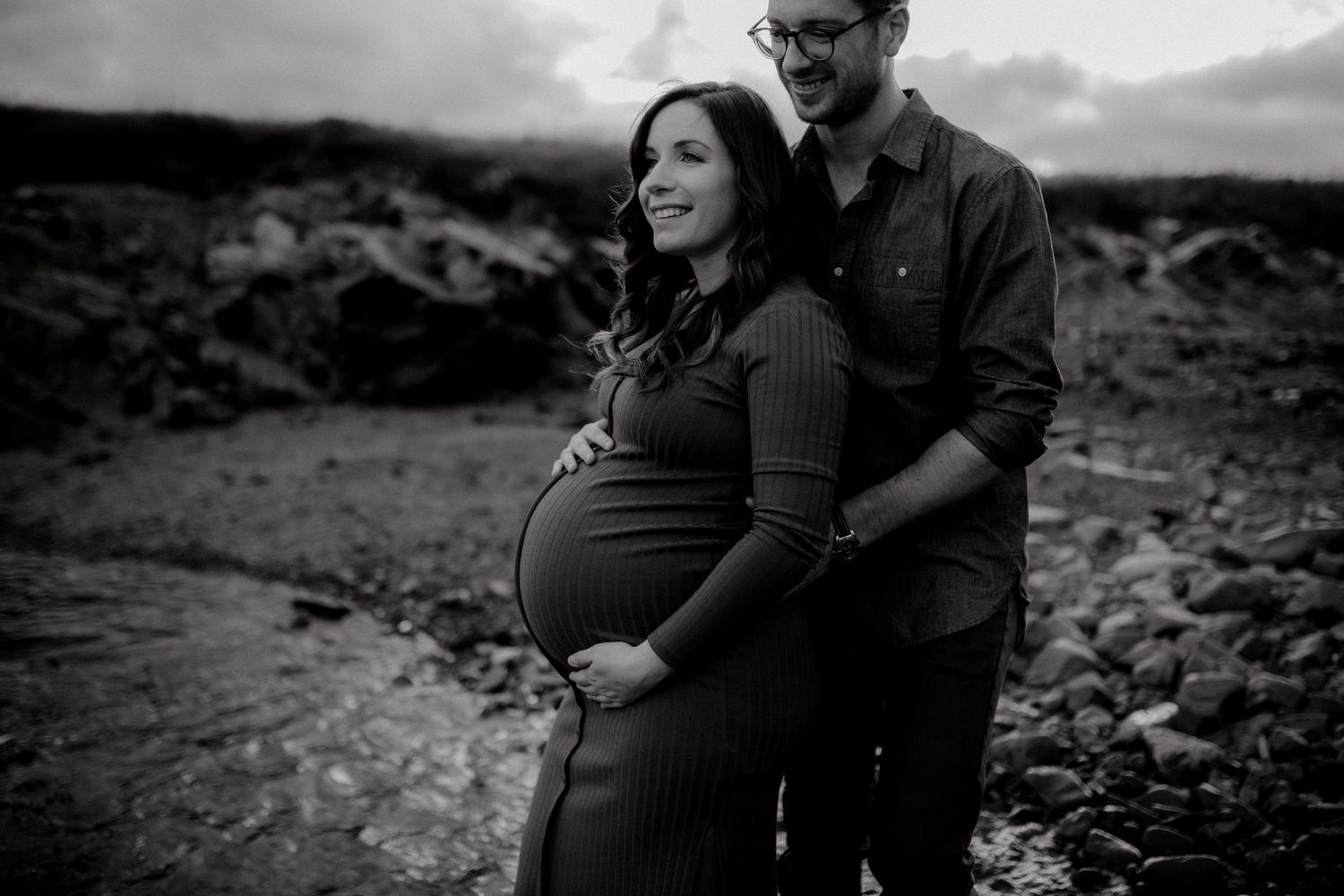 An expecting couple shares an intimate moment on a rocky beach landscape.